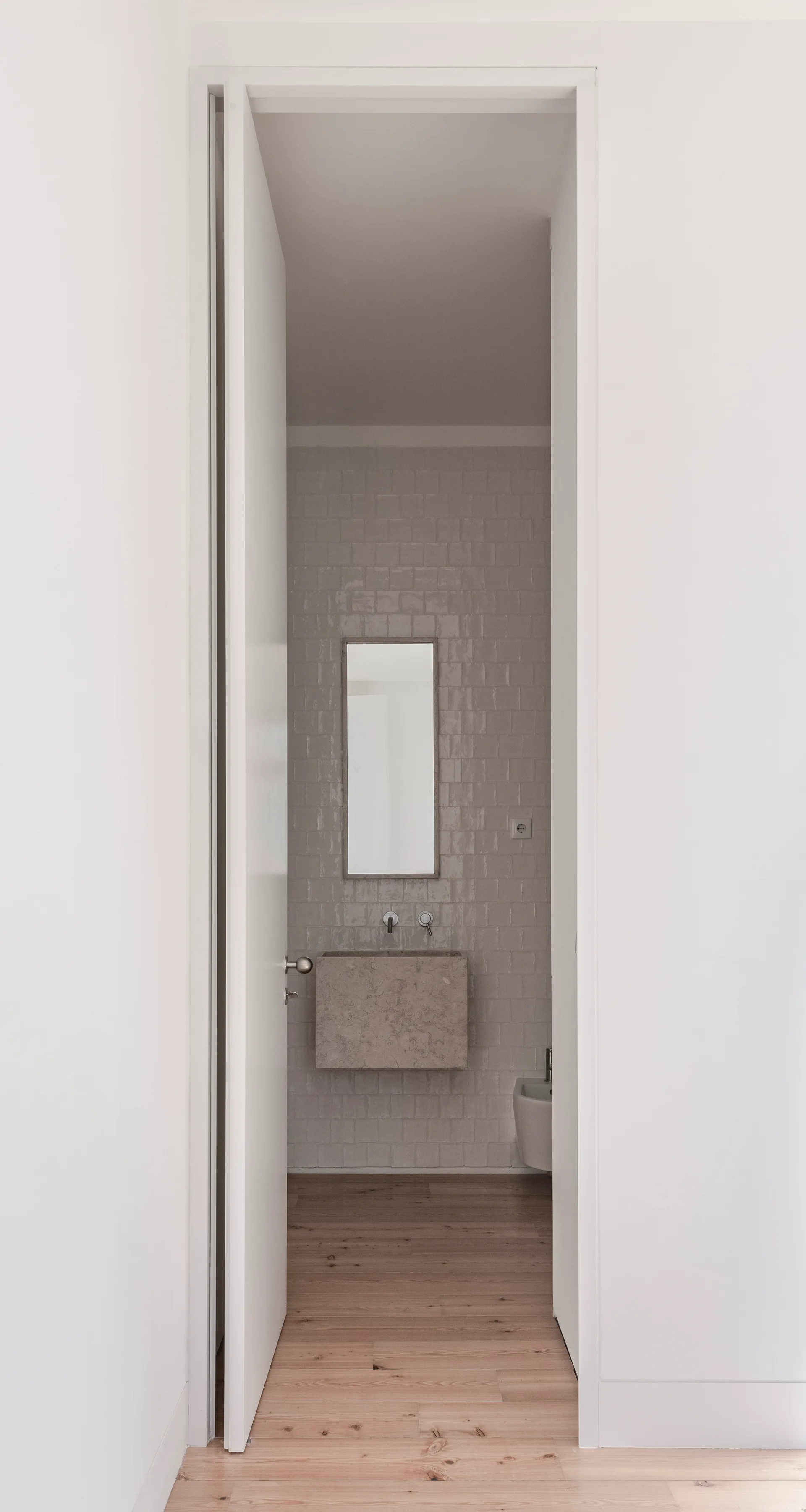 An open white door reveals a minimalist bathroom featuring a floating stone sink, a thin rectangular mirror, and glossy square wall tiles.