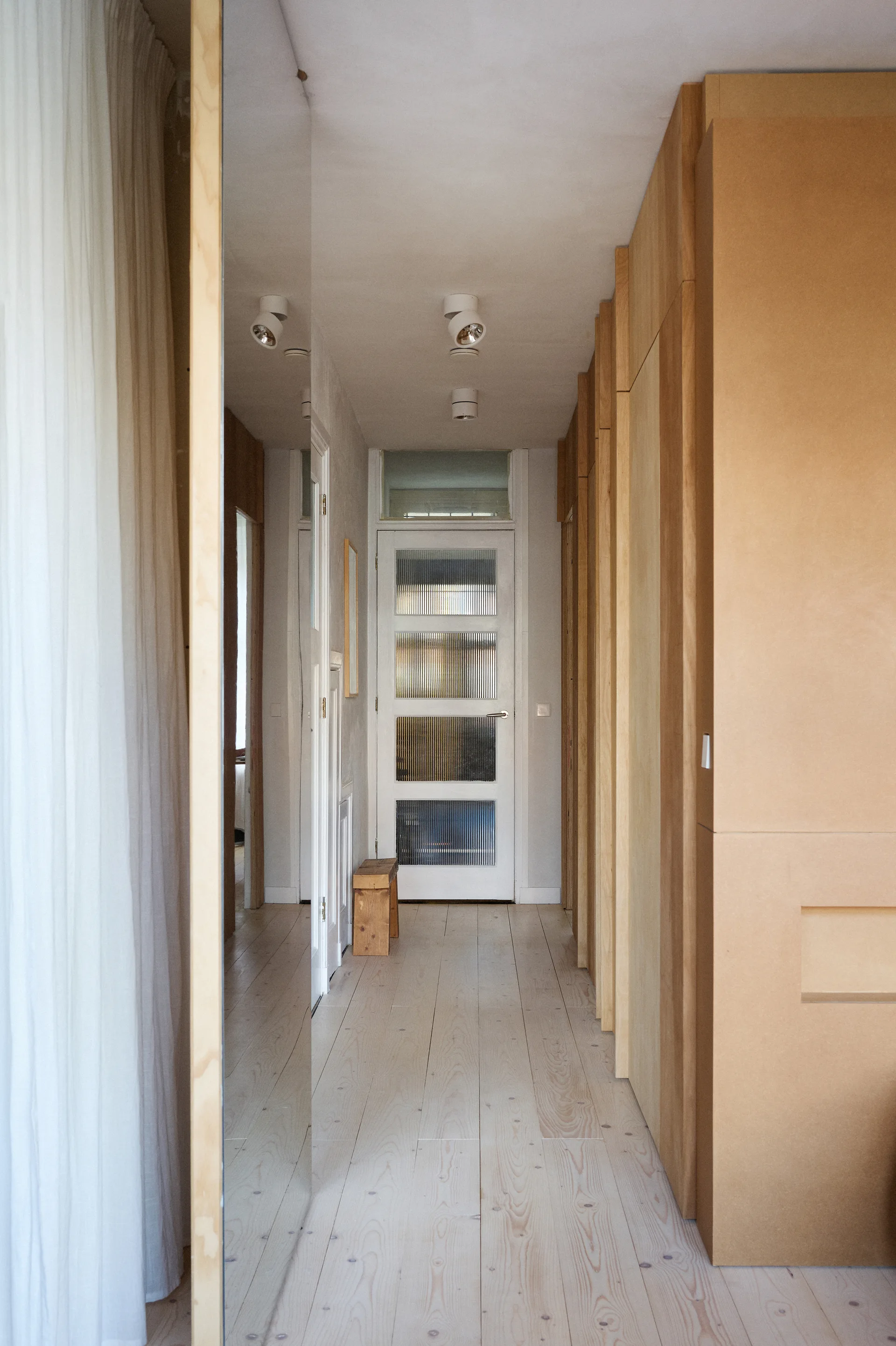 A long interior hallway features light wood floorboards, a full-height wood storage wall, and a white ribbed glass door at the far end.