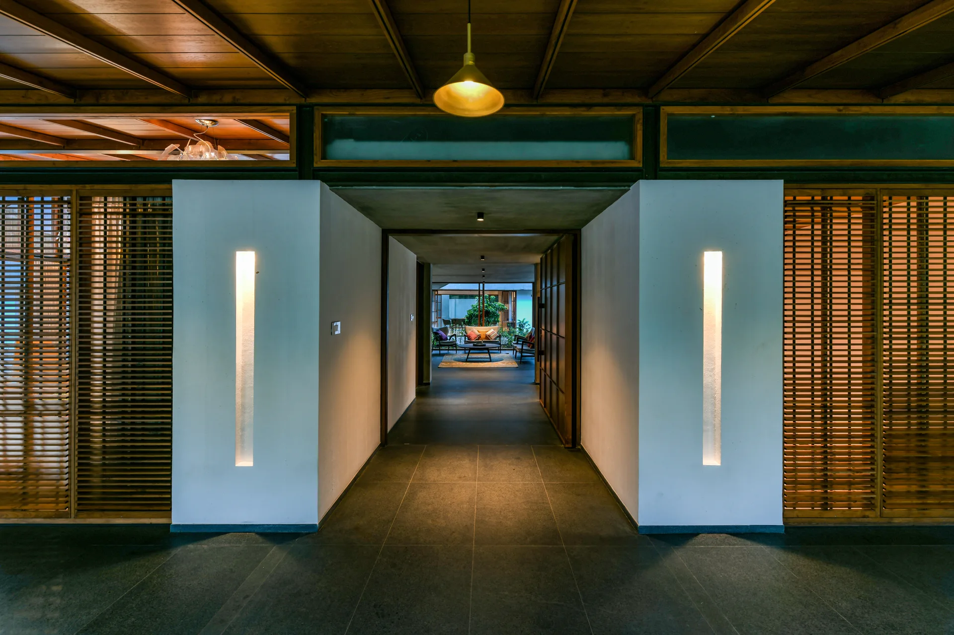 An entryway features a dark tile floor, white columns with vertical recessed lights, and warm wood slatted screens under an exposed beam ceiling.