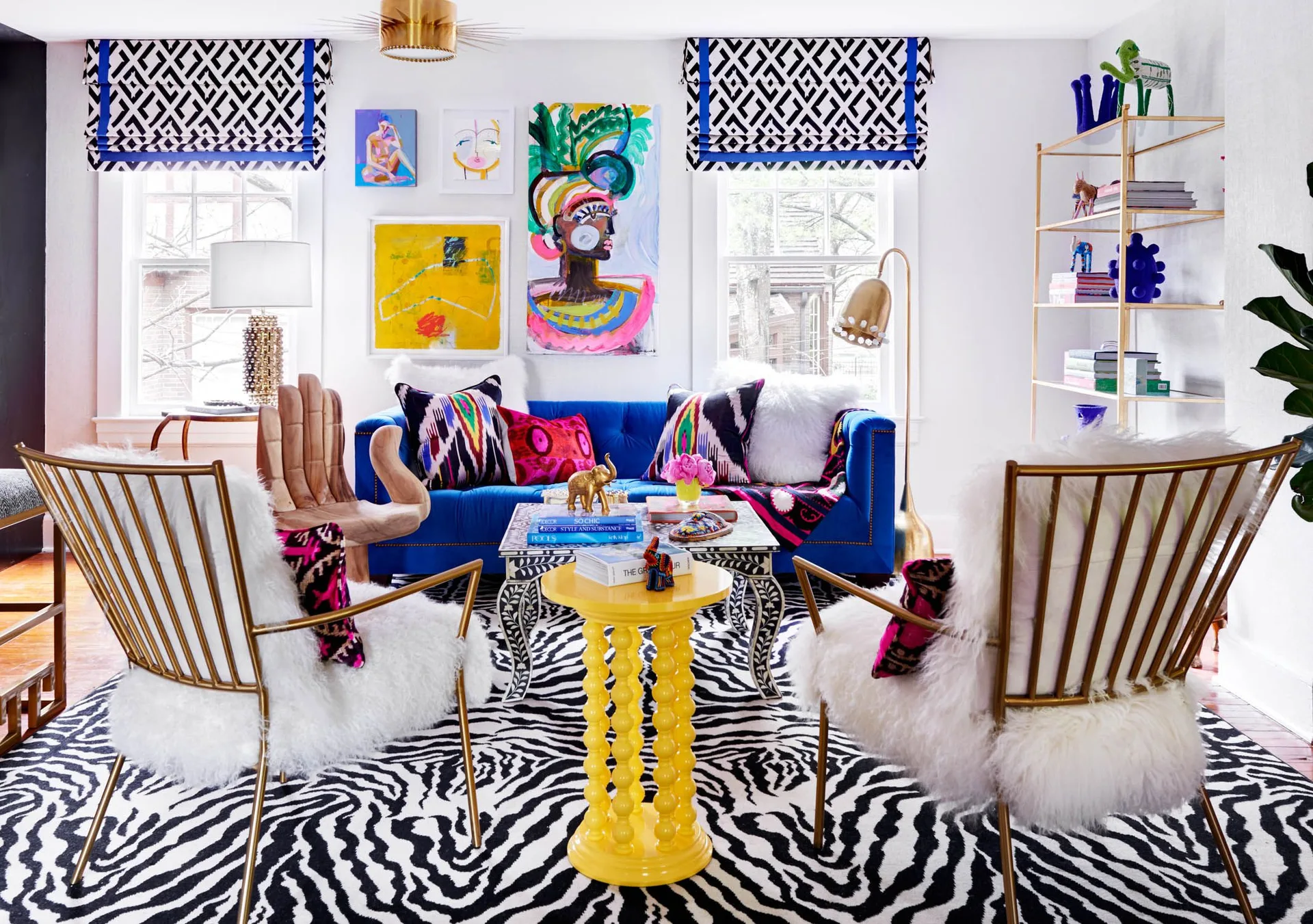 An eclectic living room with a blue velvet sofa, furry white armchairs, a black and white zebra rug, and a yellow pedestal table.