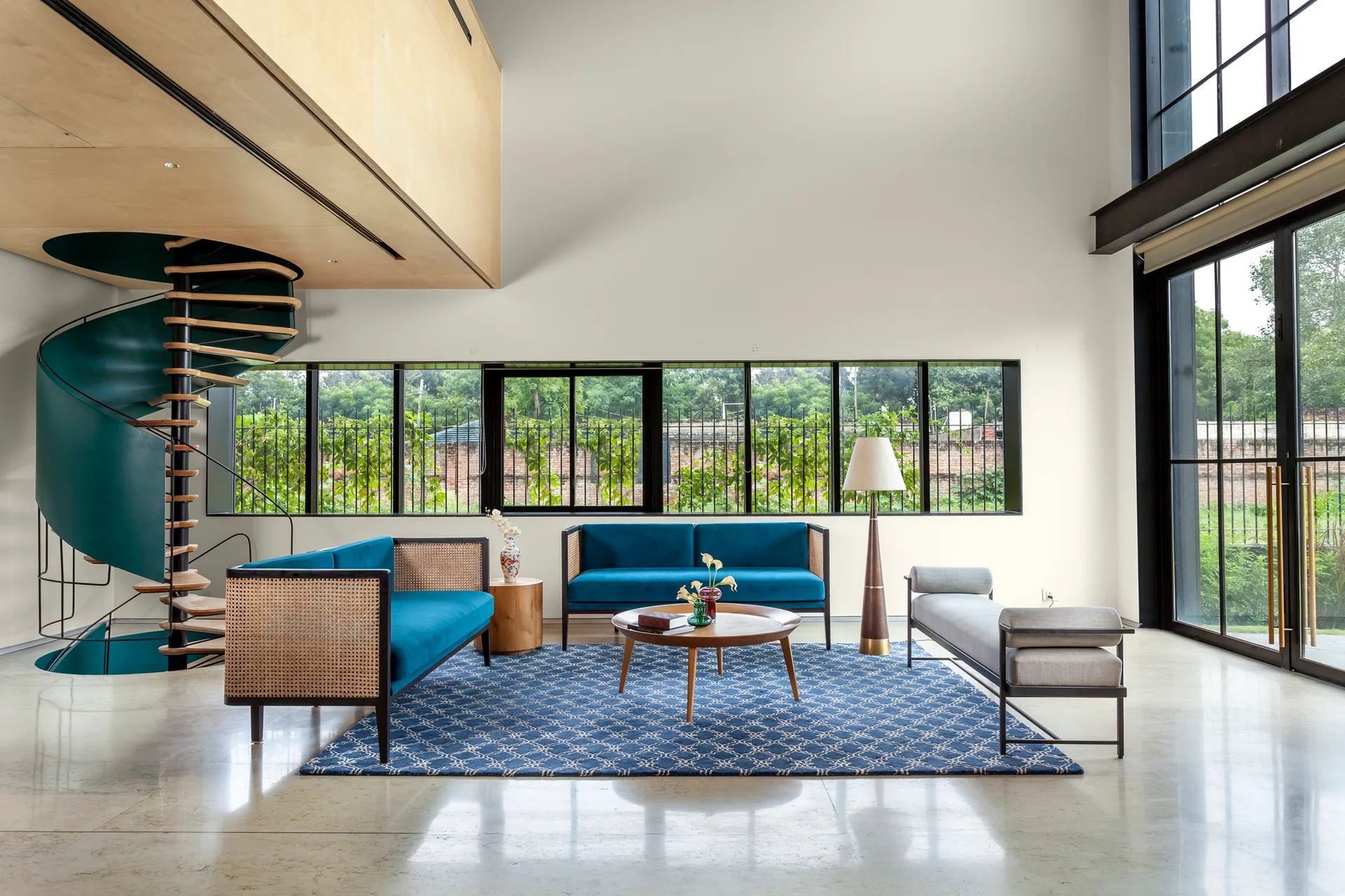 A spacious double-height living room featuring blue velvet cane-sided sofas, a blue patterned rug, and a teal spiral staircase leading to a wooden loft.