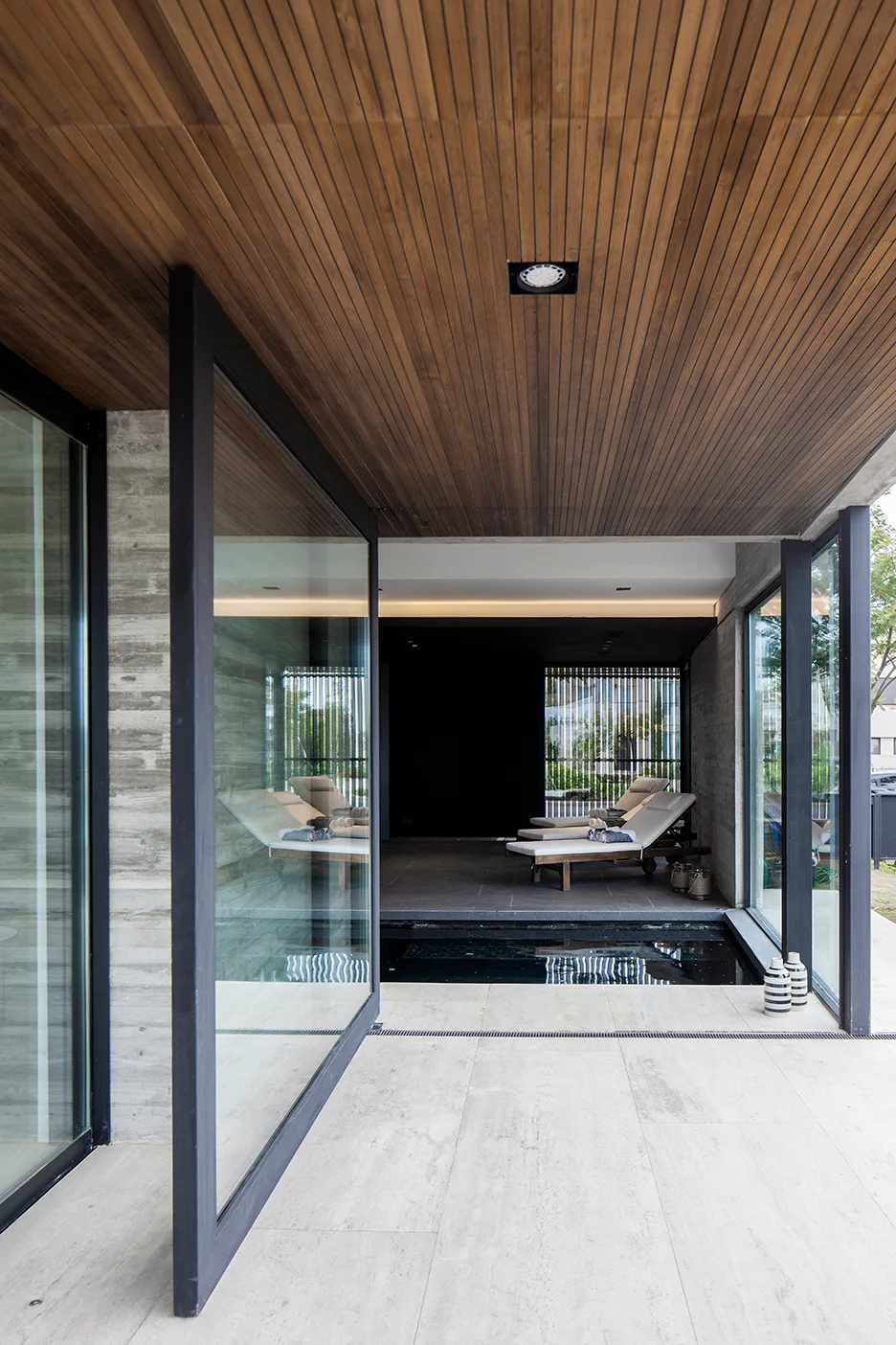 A modern indoor-outdoor patio featuring a slatted wood ceiling, a large glass pivot door, textured concrete walls, and pool lounge chairs.