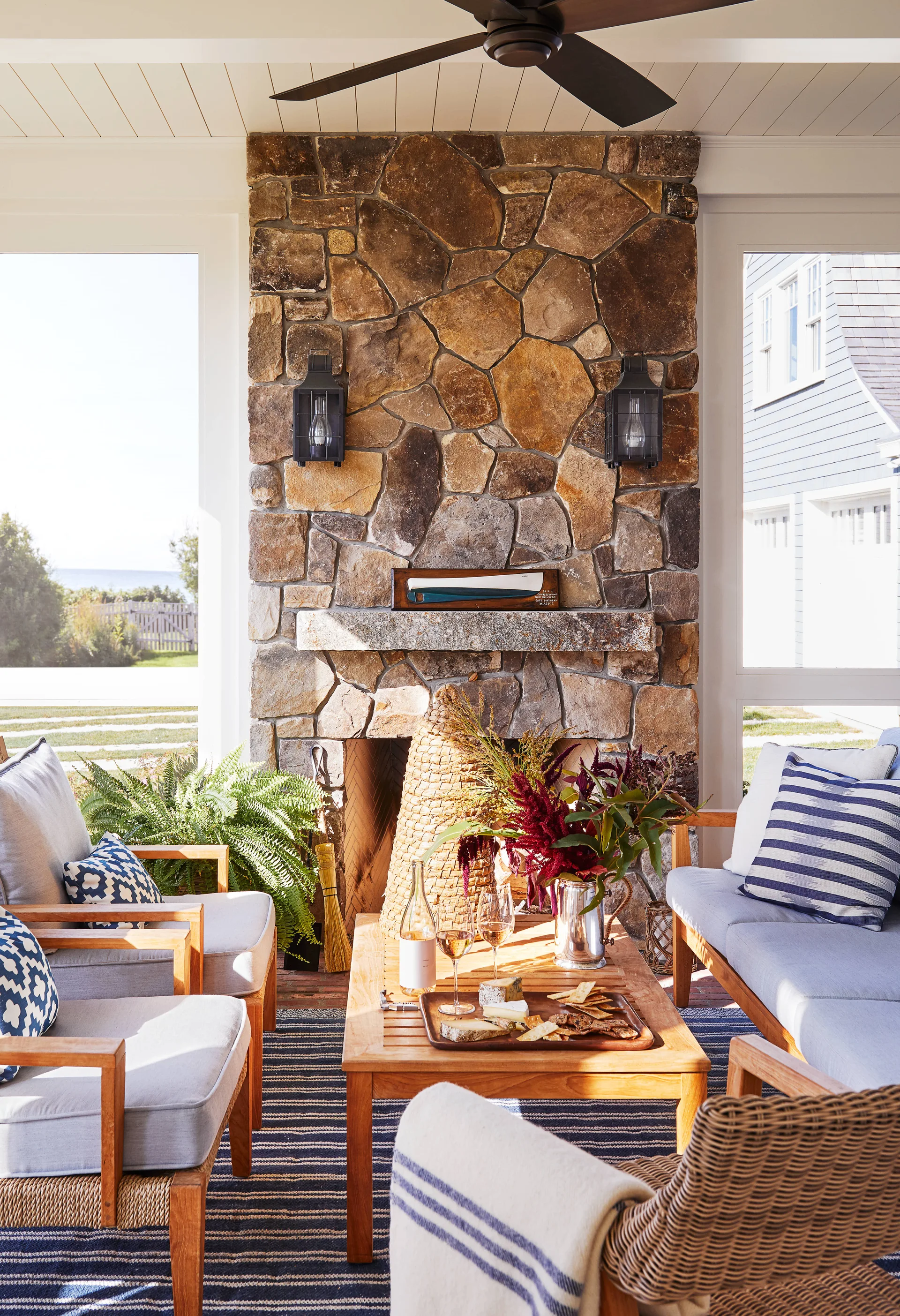 A covered porch features a tall fieldstone fireplace flanked by windows, a wood coffee table, and cushioned chairs over a blue striped rug.