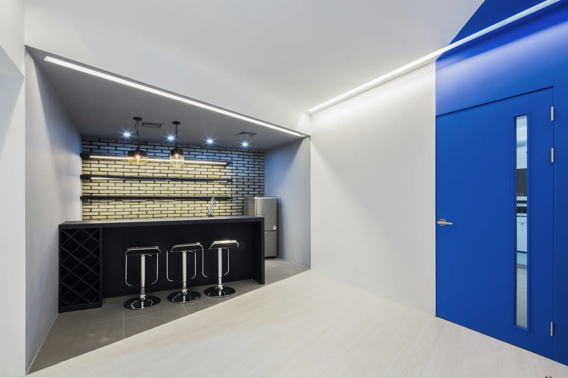 A modern home bar features a black counter with stools against a grey brick wall, positioned near a bright blue door and a diagonally split floor.