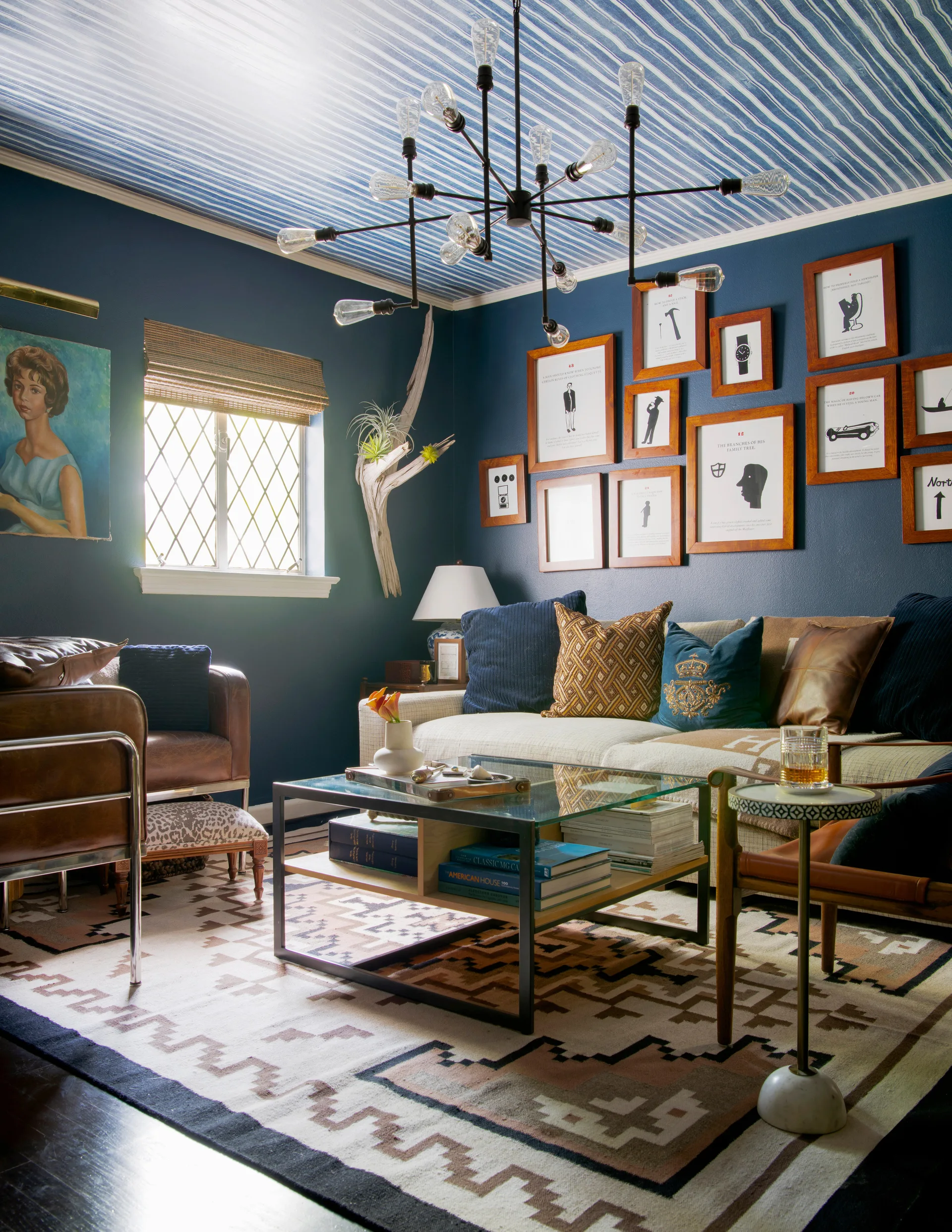 A cozy living room with deep blue walls, a striped ceiling, a glass coffee table, and a gallery wall of framed prints.