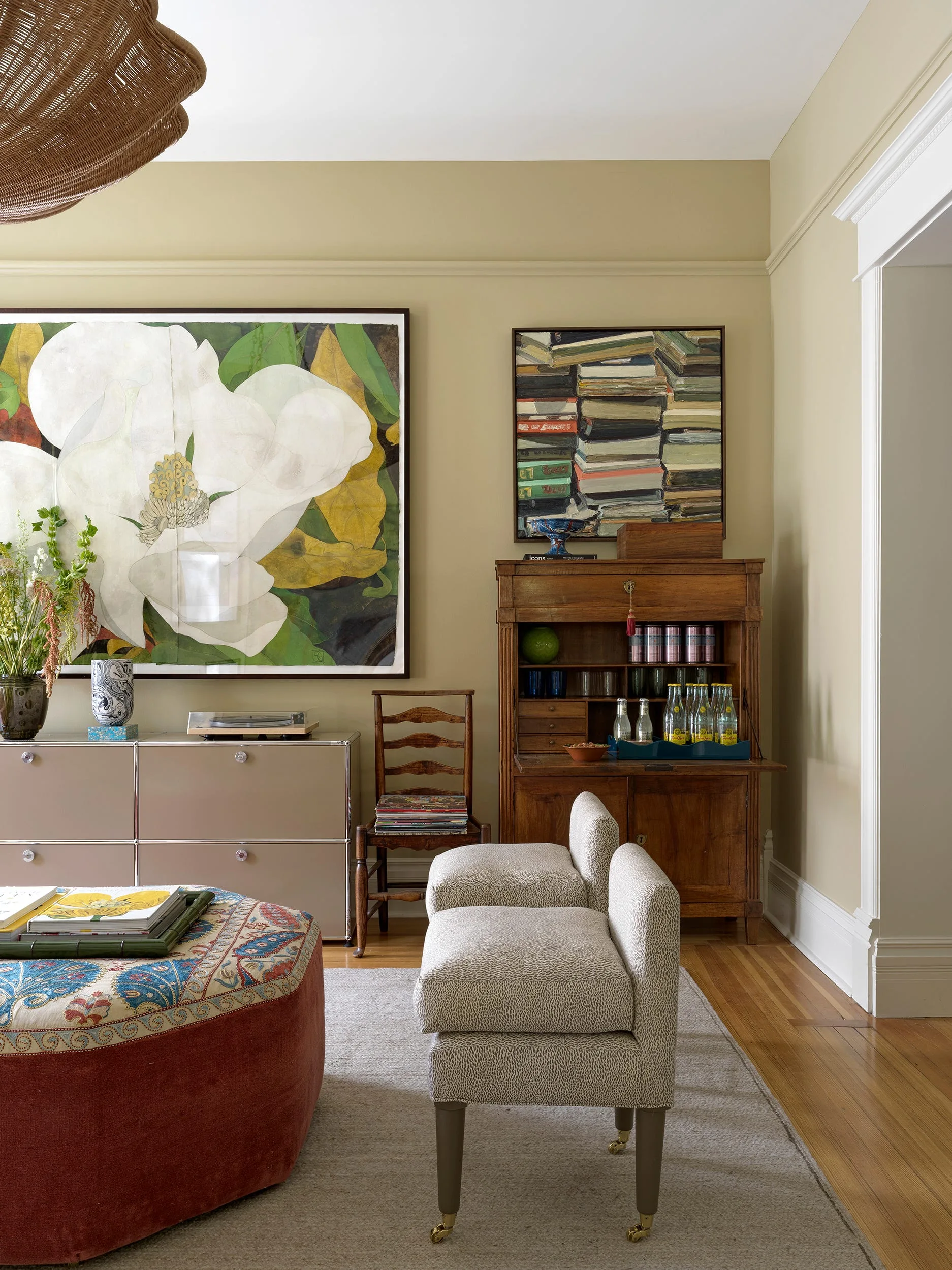 An eclectic living room with warm beige walls, a modular metal sideboard, a vintage wooden secretary, and a large patterned ottoman.