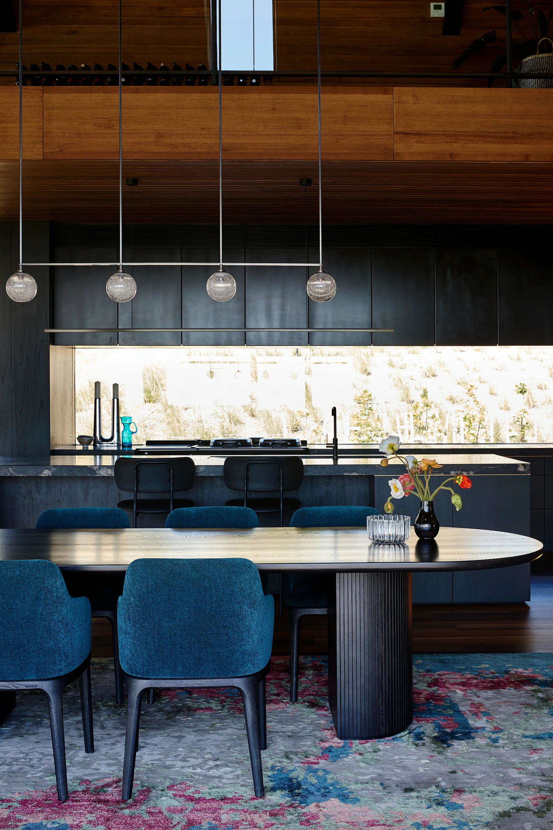 A modern dining room features textured blue chairs around a black wood table, with a dark kitchen and wood ceiling in the background.