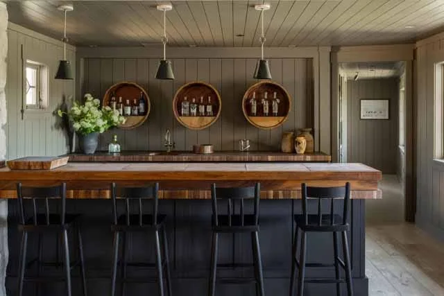 A rustic home bar featuring vertical wood wall paneling, three circular recessed display shelves, four black bar stools, and hanging pendant lights.