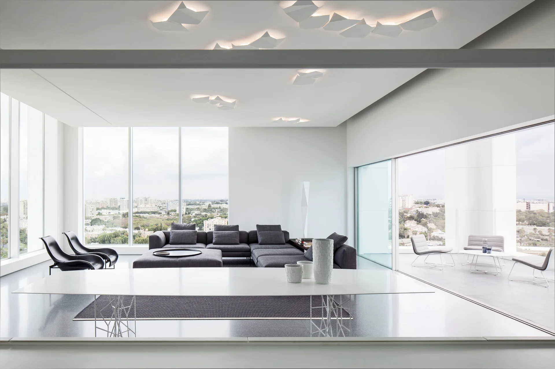 A minimalist living room features a dark grey sectional sofa, a white modern dining table, and floor-to-ceiling windows showing a city view.