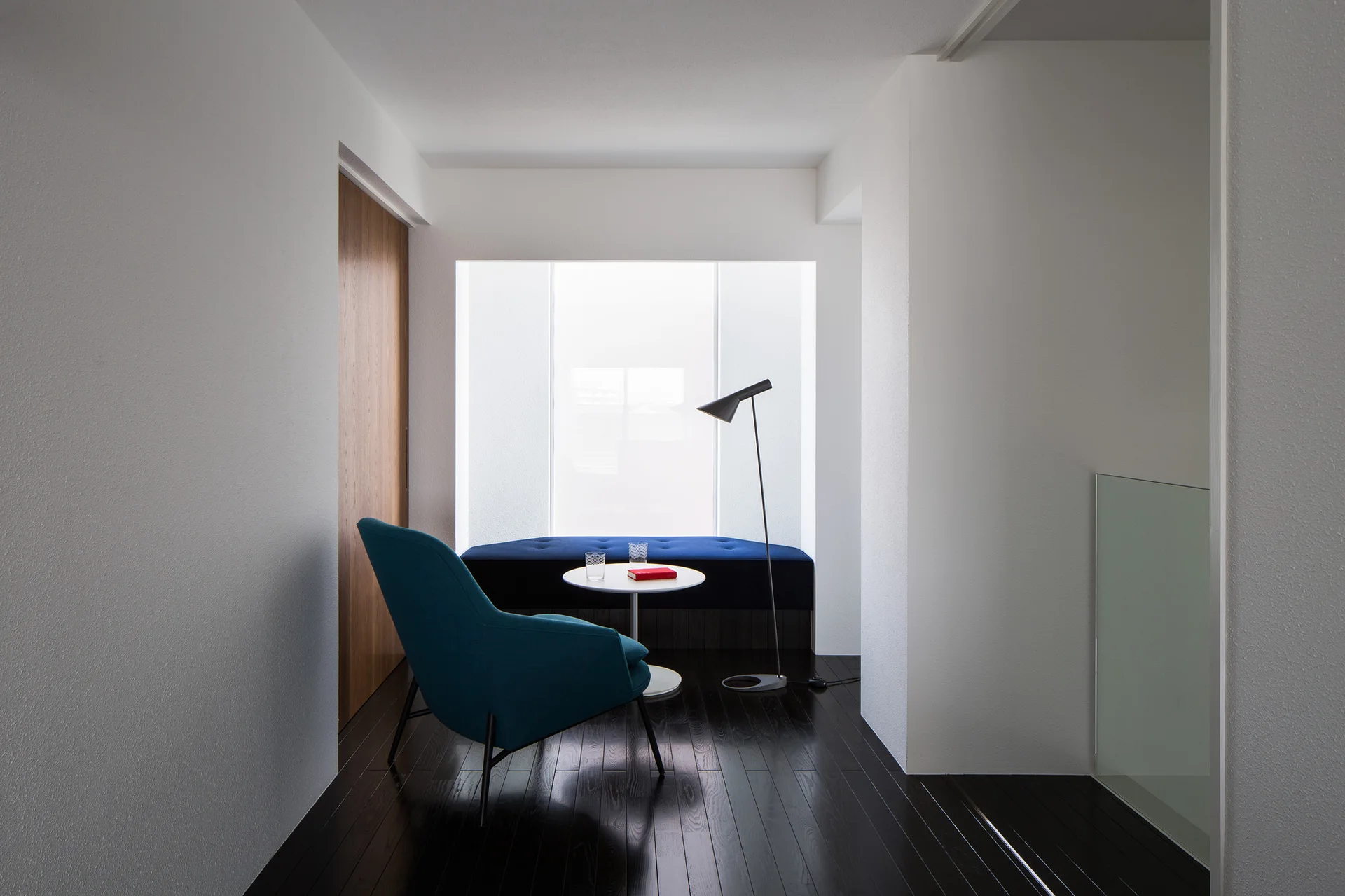 A minimalist seating area features a teal armchair and a navy upholstered bench set in a bright window nook over dark wood floors.