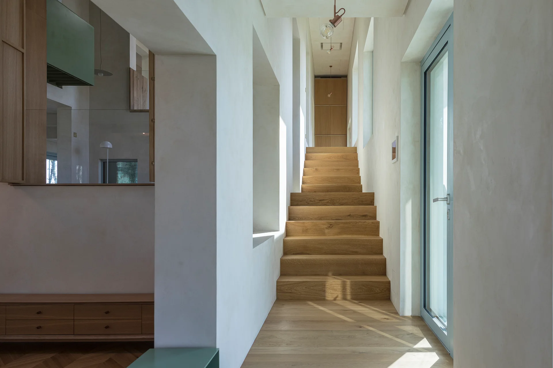 A minimalist hallway features a central light-toned wooden staircase framed by smooth plaster walls and illuminated by natural light.