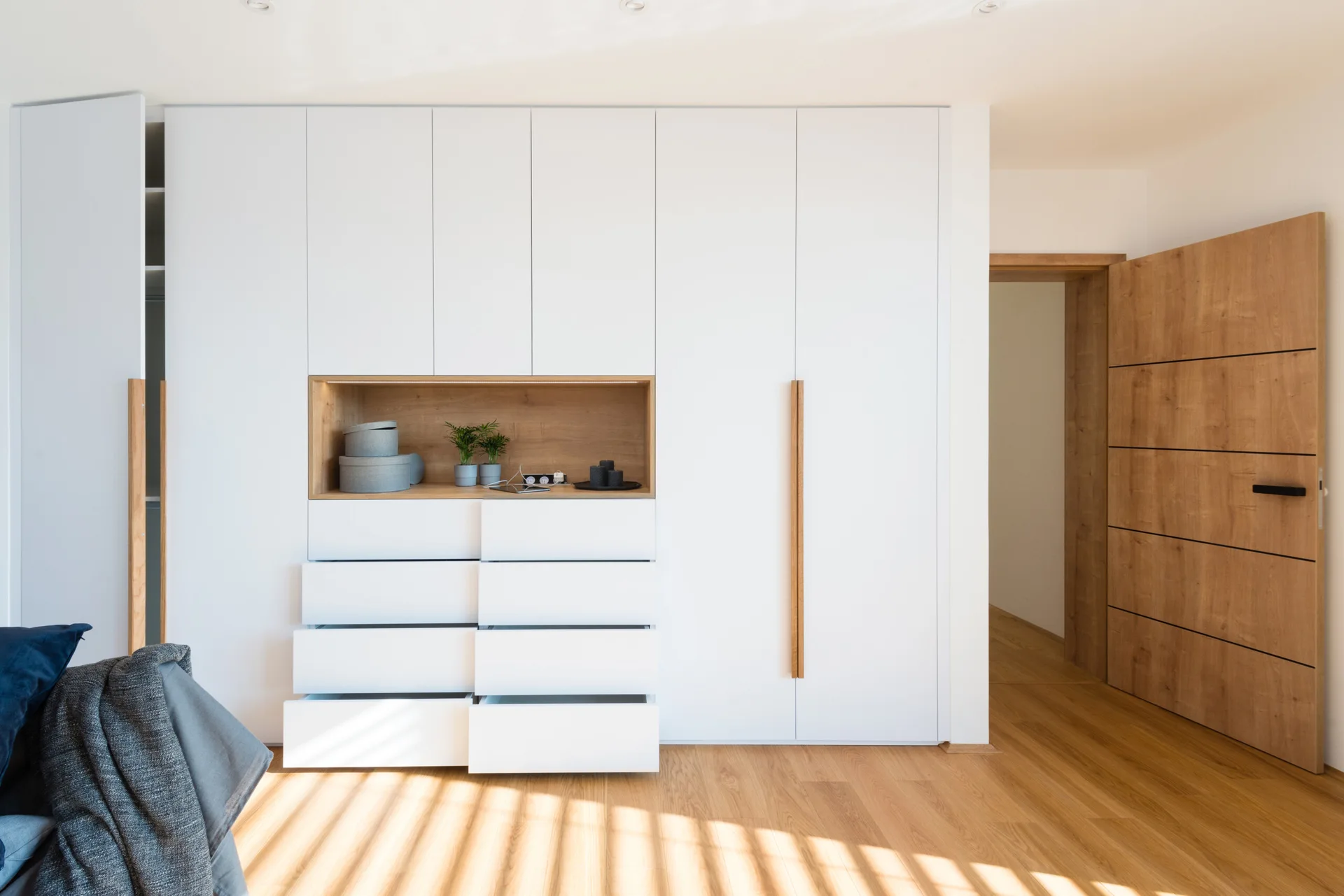 A modern floor-to-ceiling white wardrobe features a wood-lined display niche and lower drawers on light wood flooring.