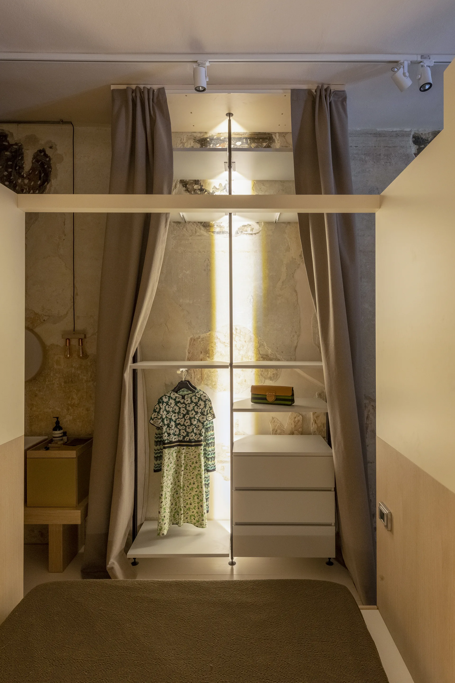 A minimalist open closet featuring white shelves and drawers framed by taupe curtains against a distressed concrete wall under track lighting.