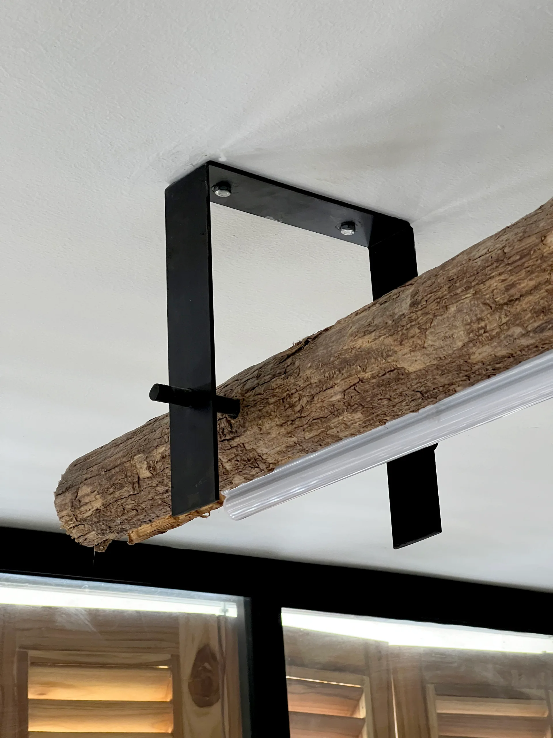 A rough natural log ceiling beam is suspended from a white ceiling by a matte black U-shaped metal bracket.