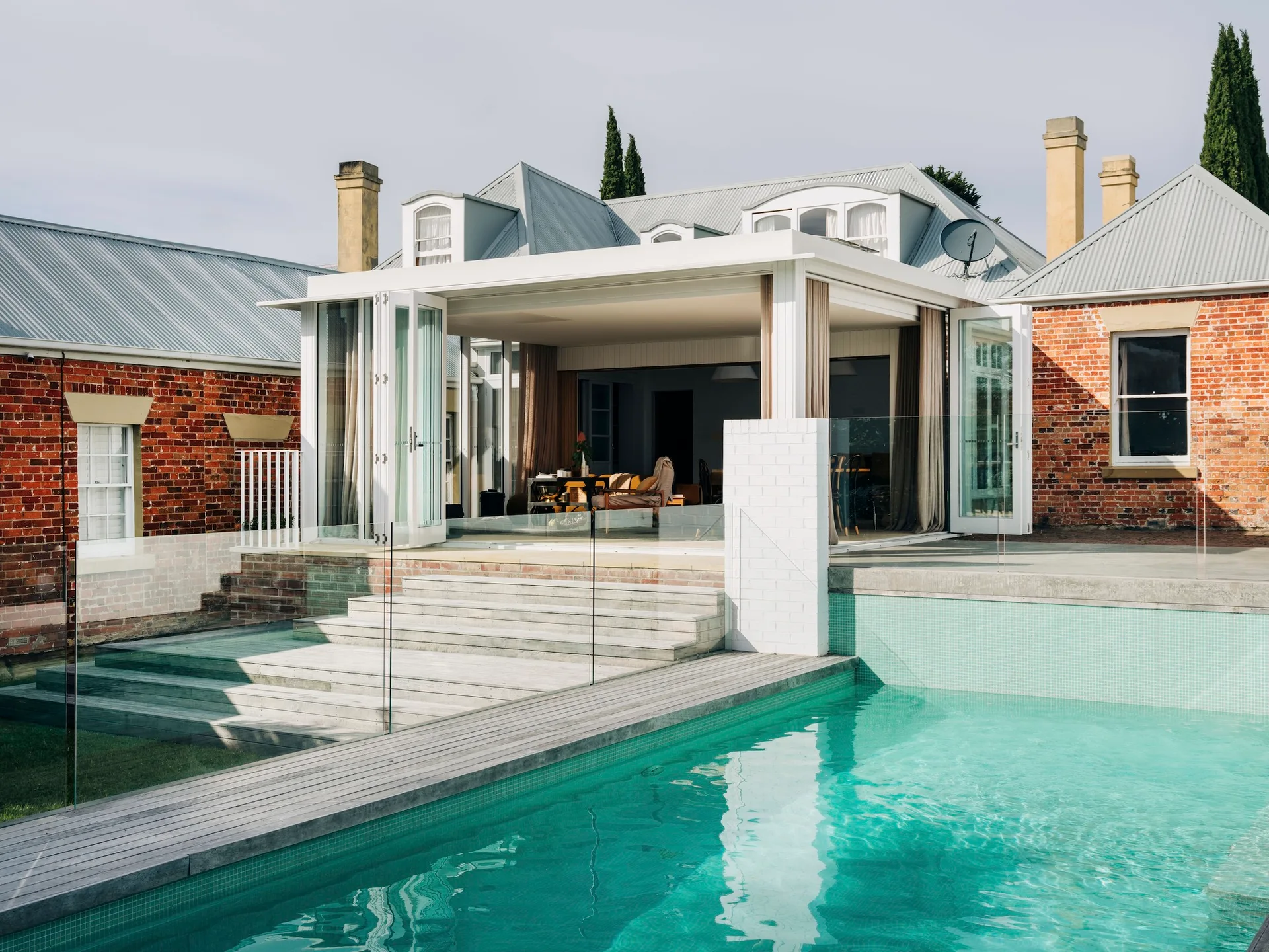 A rectangular swimming pool with a wood deck and glass railing sits in front of a covered patio and red brick residential house.