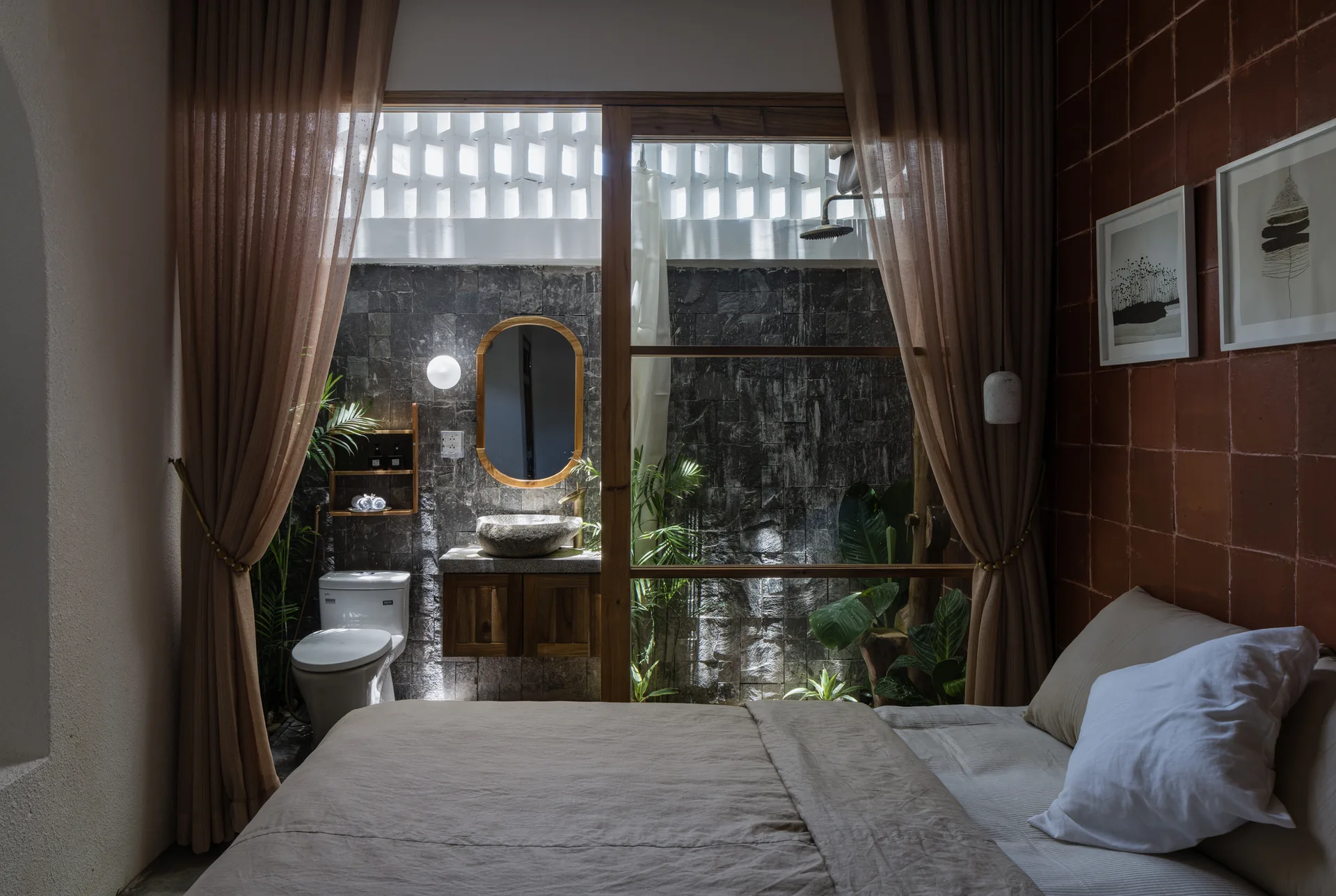 A bedroom with beige bedding features a sliding glass door looking into an integrated bathroom with a dark stone wall, wood vanity, and plants.