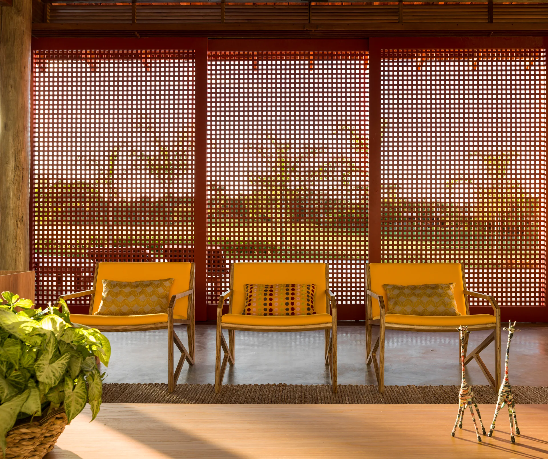 Three yellow armchairs with patterned pillows stand side-by-side in front of a tall red geometric lattice screen in a sunlit room.