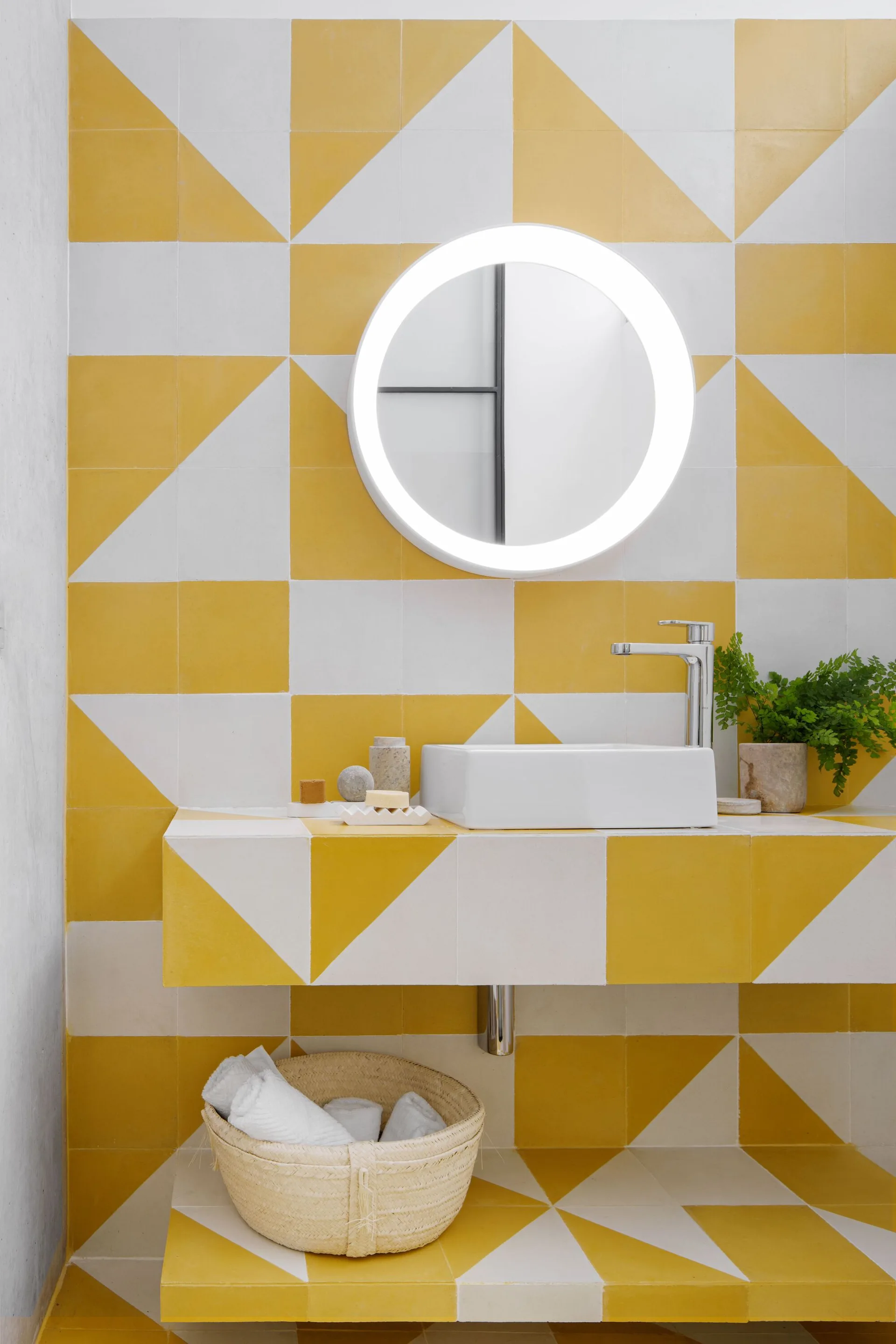 A modern bathroom vanity featuring a vessel sink and a round backlit mirror against a yellow and white geometric tile wall.