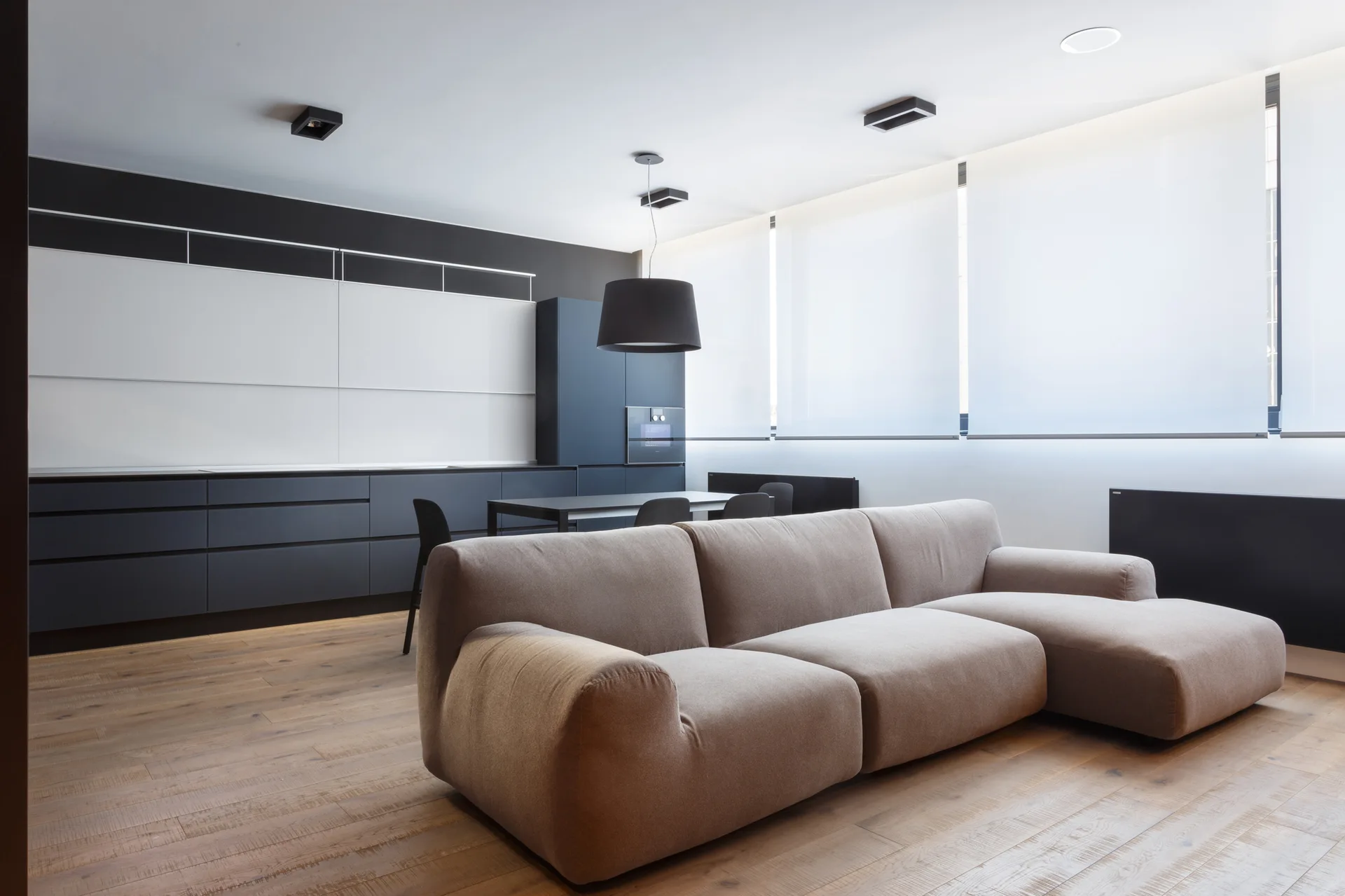 A tan modular sofa sits on light wood floors in an open-plan room facing a black minimalist kitchen and large windows with white roller shades.