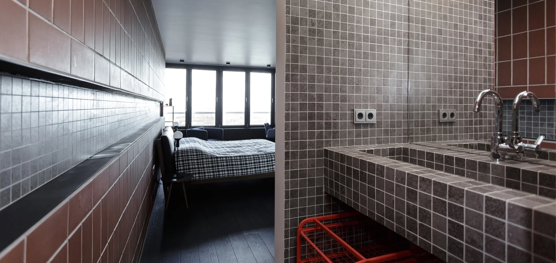 A grey mosaic tiled bathroom vanity with a chrome faucet stands next to a terracotta wall looking into a bedroom with a plaid bed.