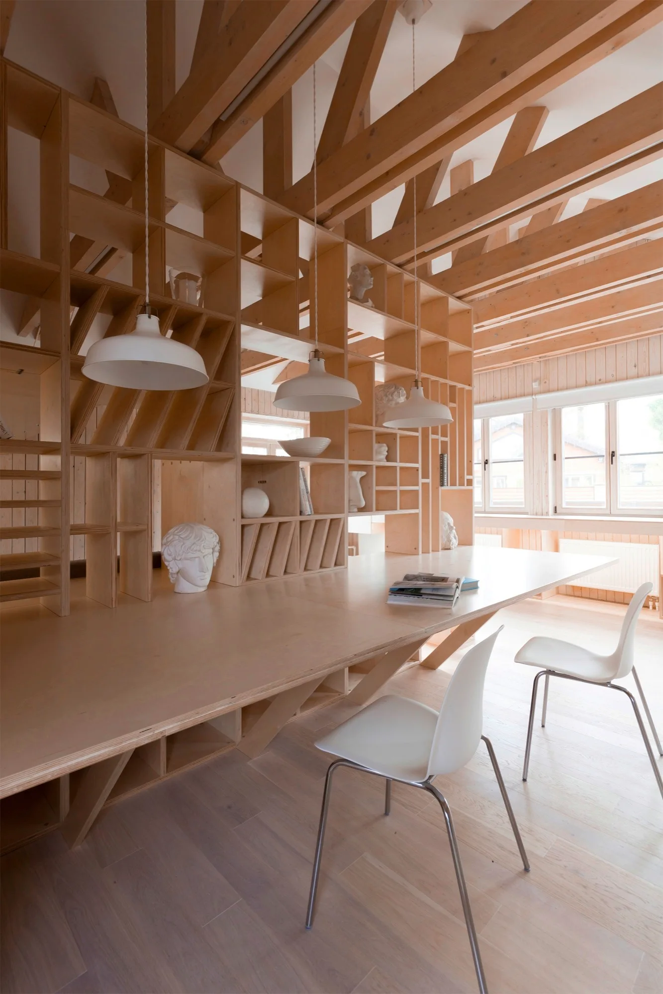 A minimalist workspace features a custom plywood desk integrated with a large shelving unit under exposed timber ceiling beams with white chairs.