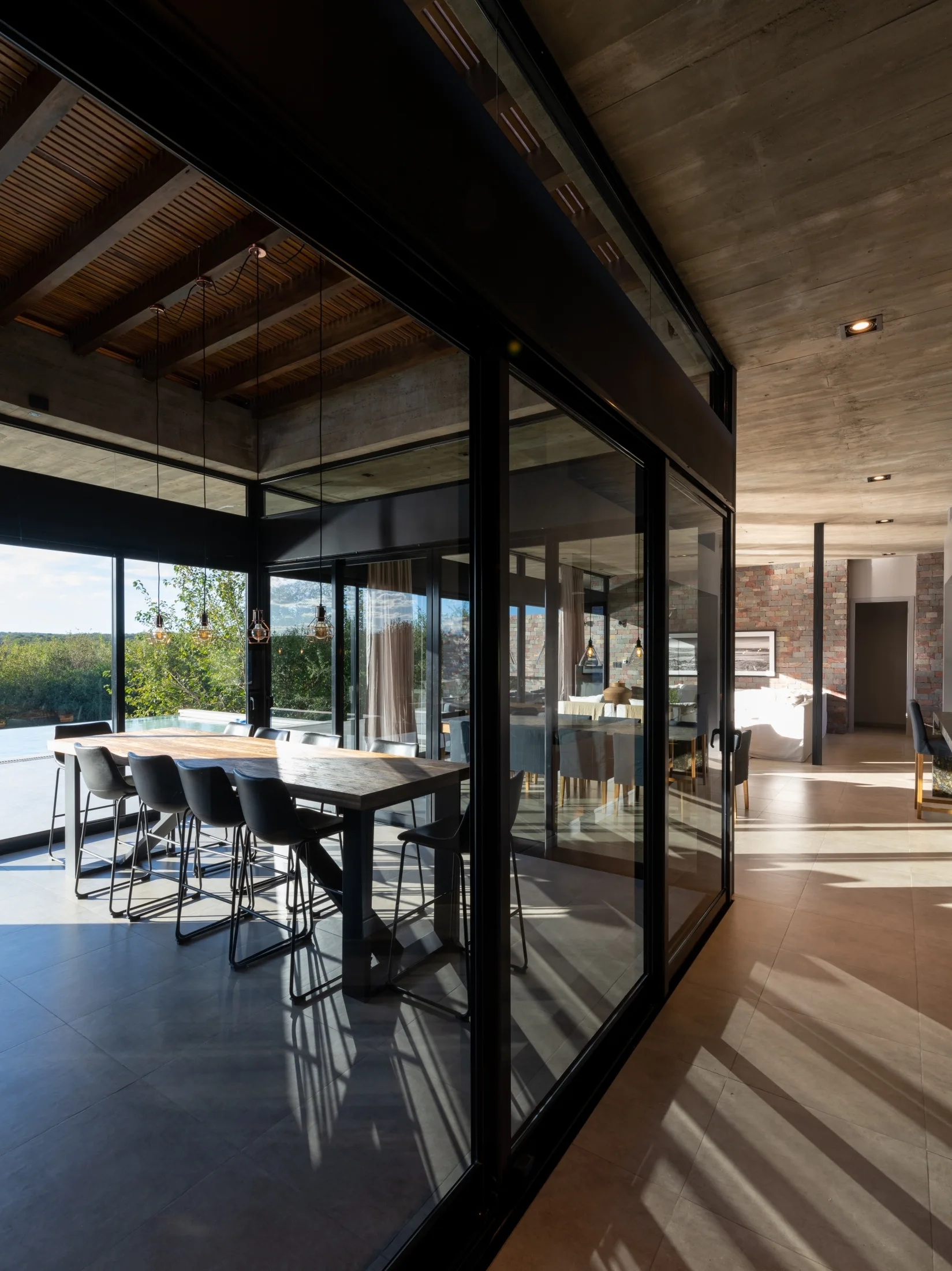 A modern dining area features a wood table, black chairs, and pendant lights enclosed by floor-to-ceiling glass walls under a concrete ceiling.