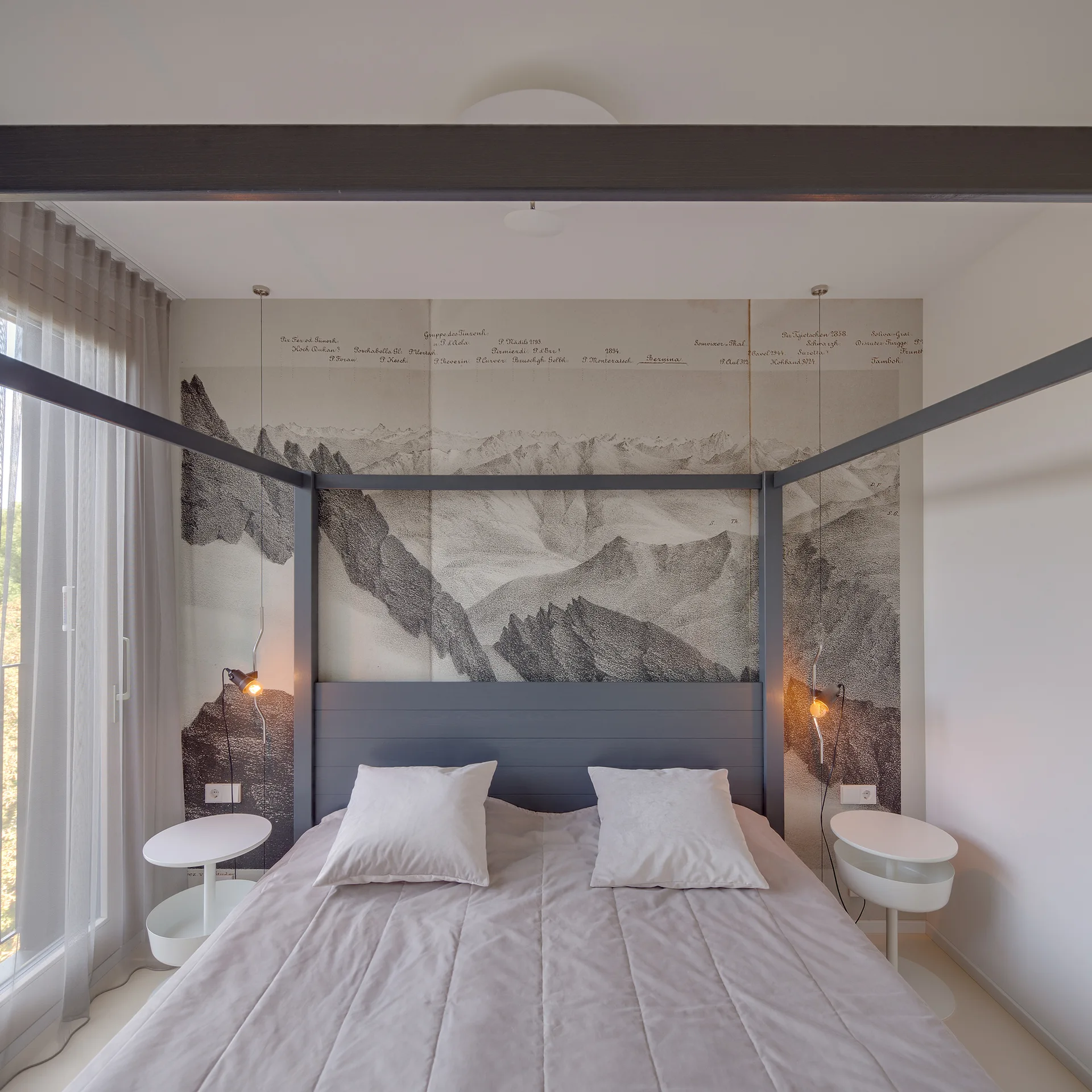 A grey minimalist canopy bed stands against a black and white mountain mural flanked by modern round white nightstands.