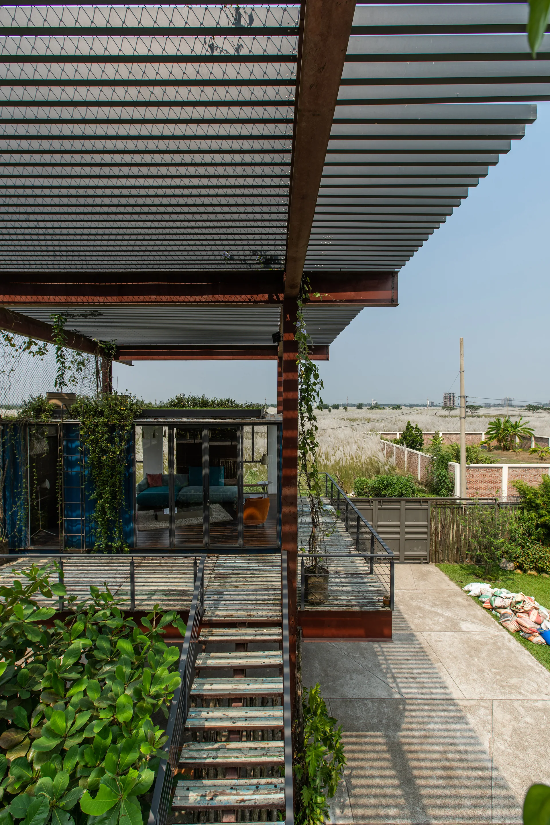 An elevated wooden deck and staircase featuring a slatted metal canopy and a blue container living space with a glass facade.