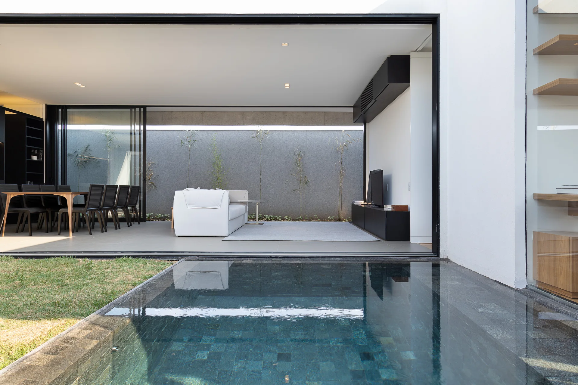 A minimalist indoor living area with a white sofa opens onto a backyard pool through a large sliding glass doorway.