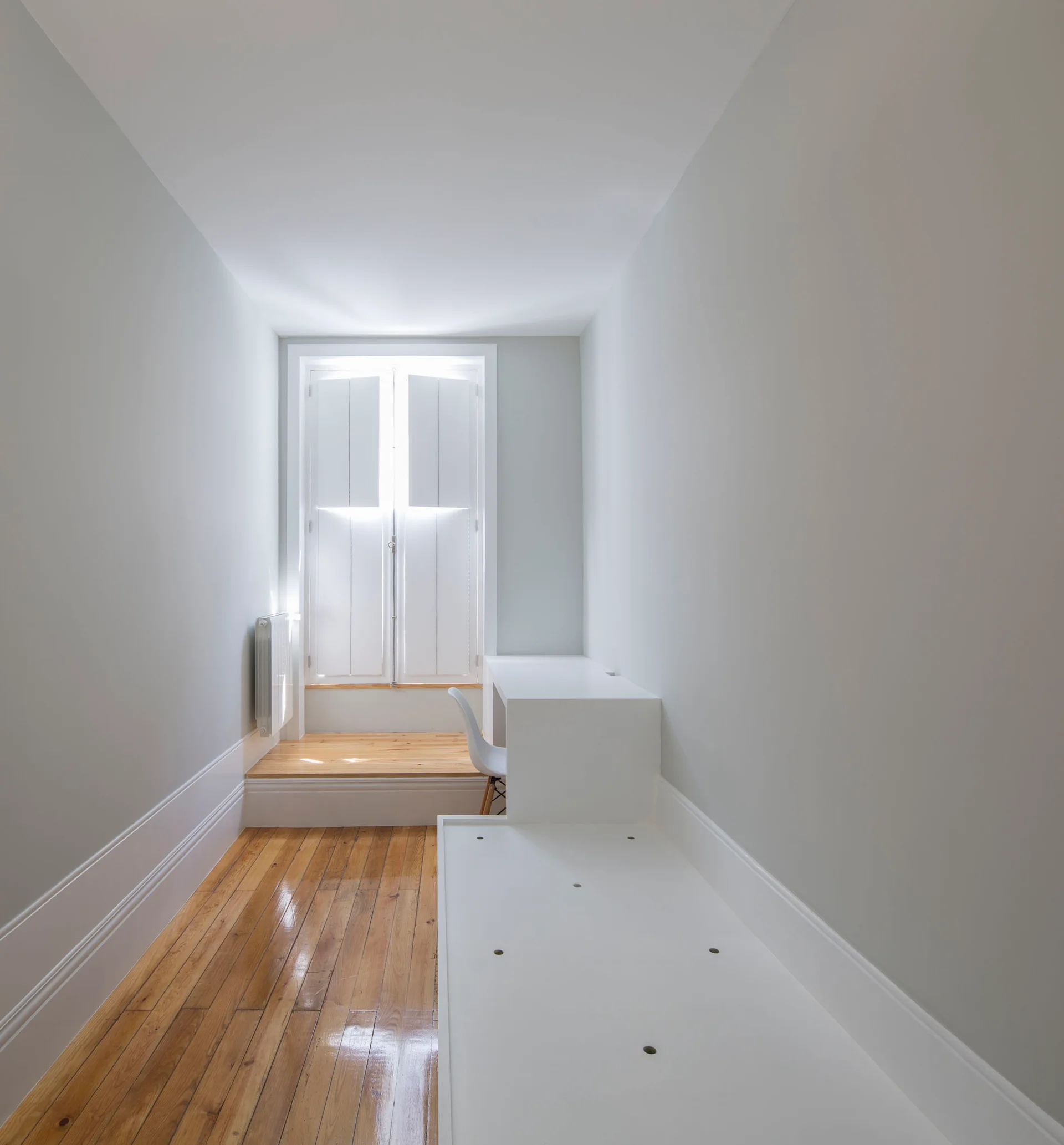 A narrow white minimalist home office featuring glossy wood floors, a built-in desk with a white chair, and white shutters on a window.