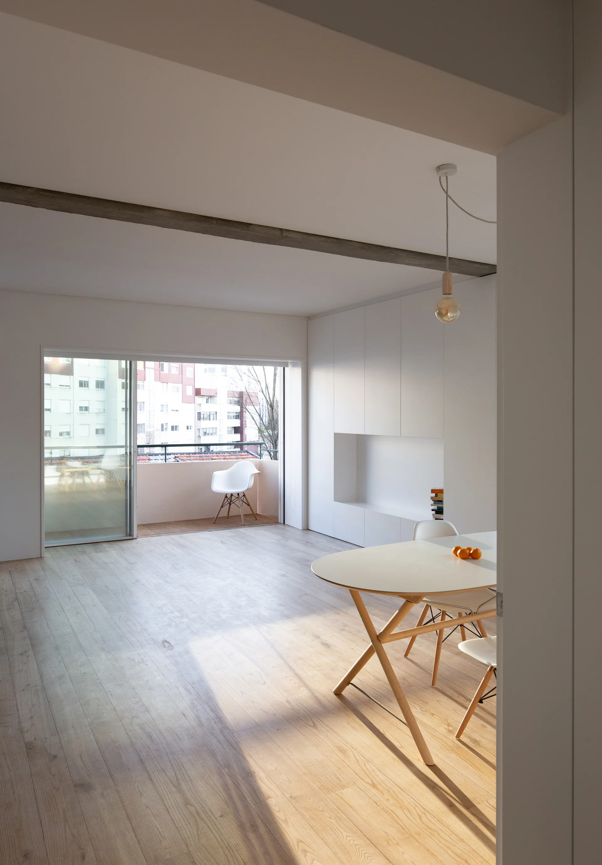 A minimalist dining area features a white oval table and chairs next to a large built-in storage wall, with a balcony visible in the background.