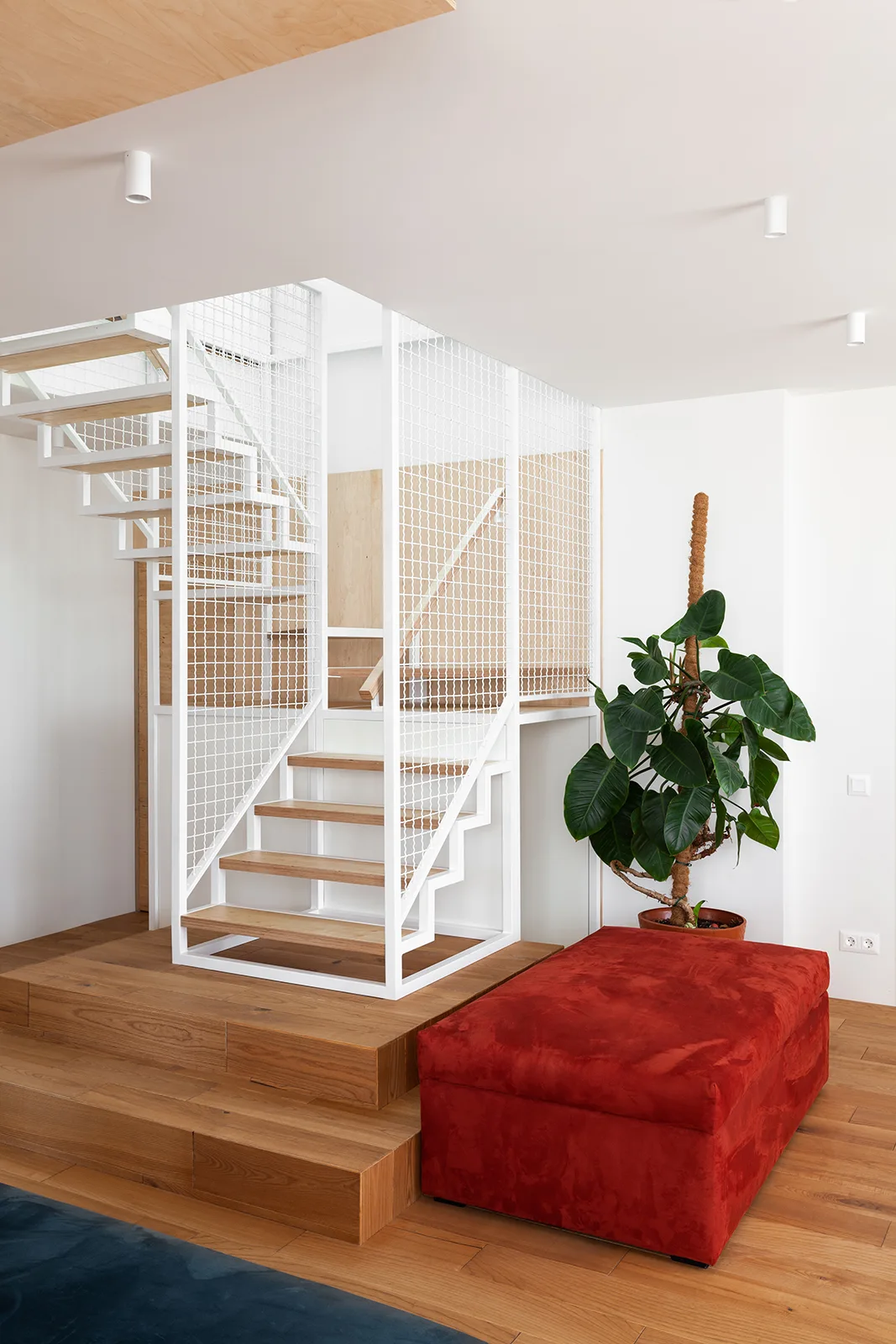A modern staircase with white metal grid railing and wooden steps rests on a tiered wood platform next to a red ottoman.