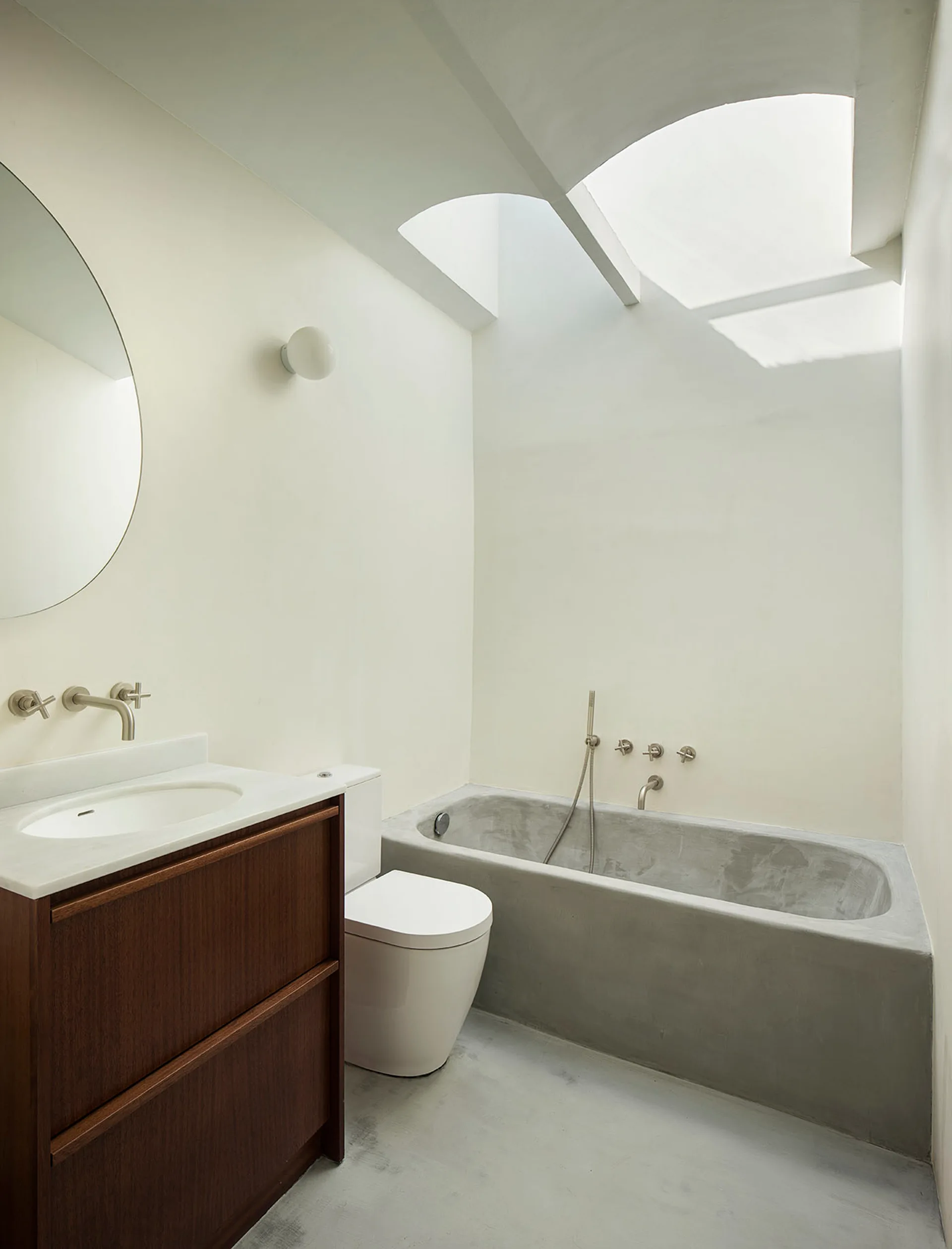 A minimalist bathroom featuring a dark wood vanity, a round mirror, a white toilet, and a concrete bathtub beneath a large skylight.