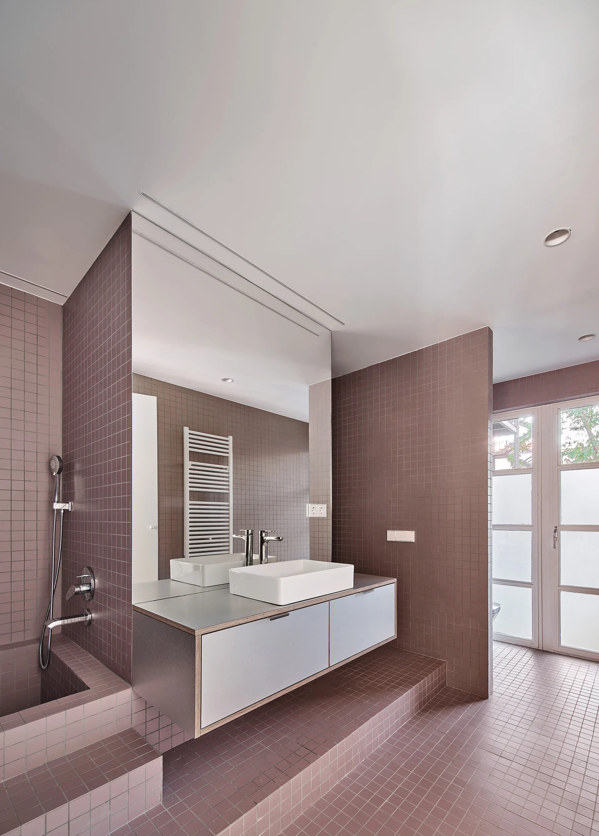 A minimalist bathroom features mauve square tiles on the walls and floors, a grey floating vanity, a white vessel sink, and a large wall mirror.