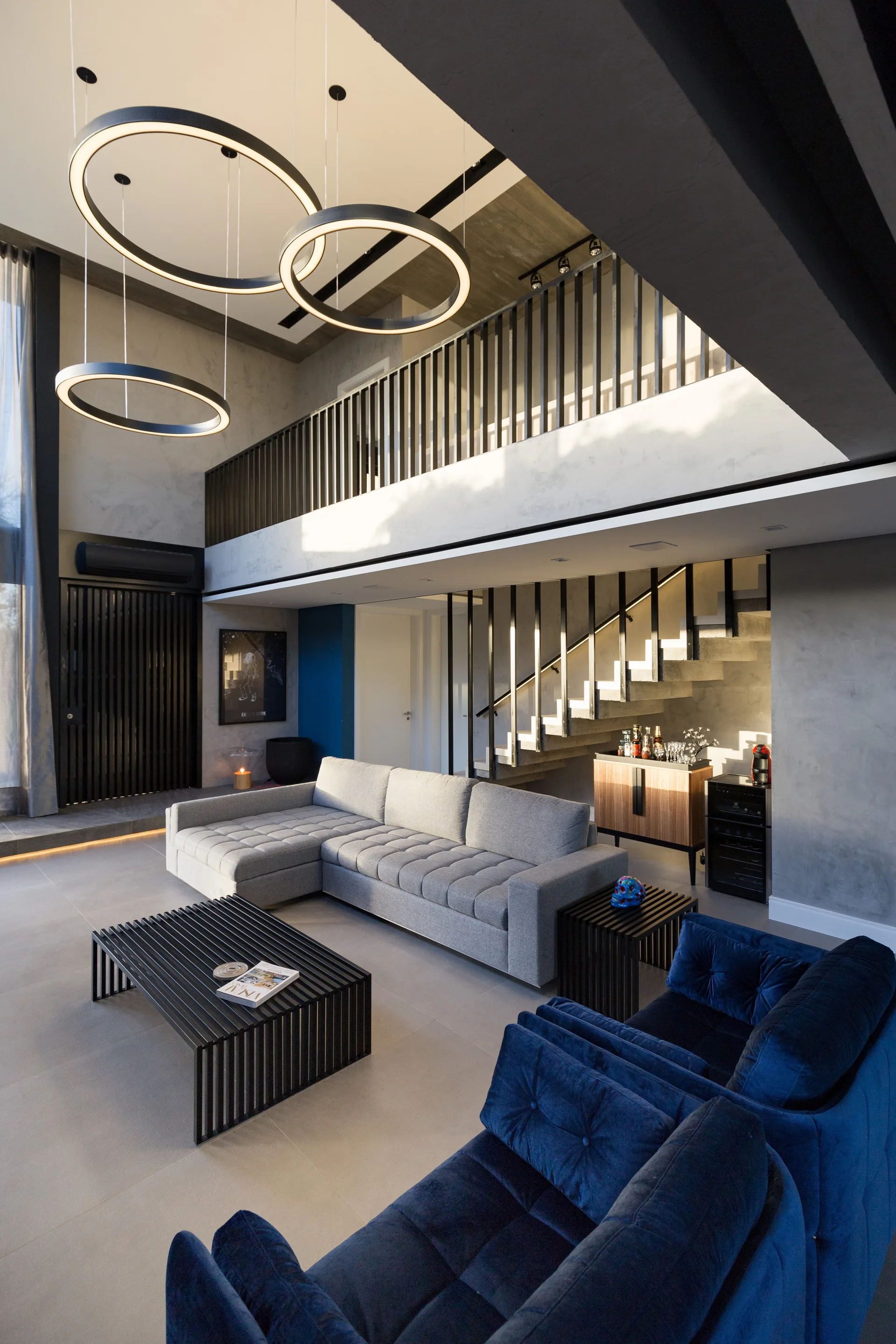 A double-height living room features a grey sectional, navy blue velvet chairs, black slatted coffee table, and large ring pendant lights below a mezzanine.
