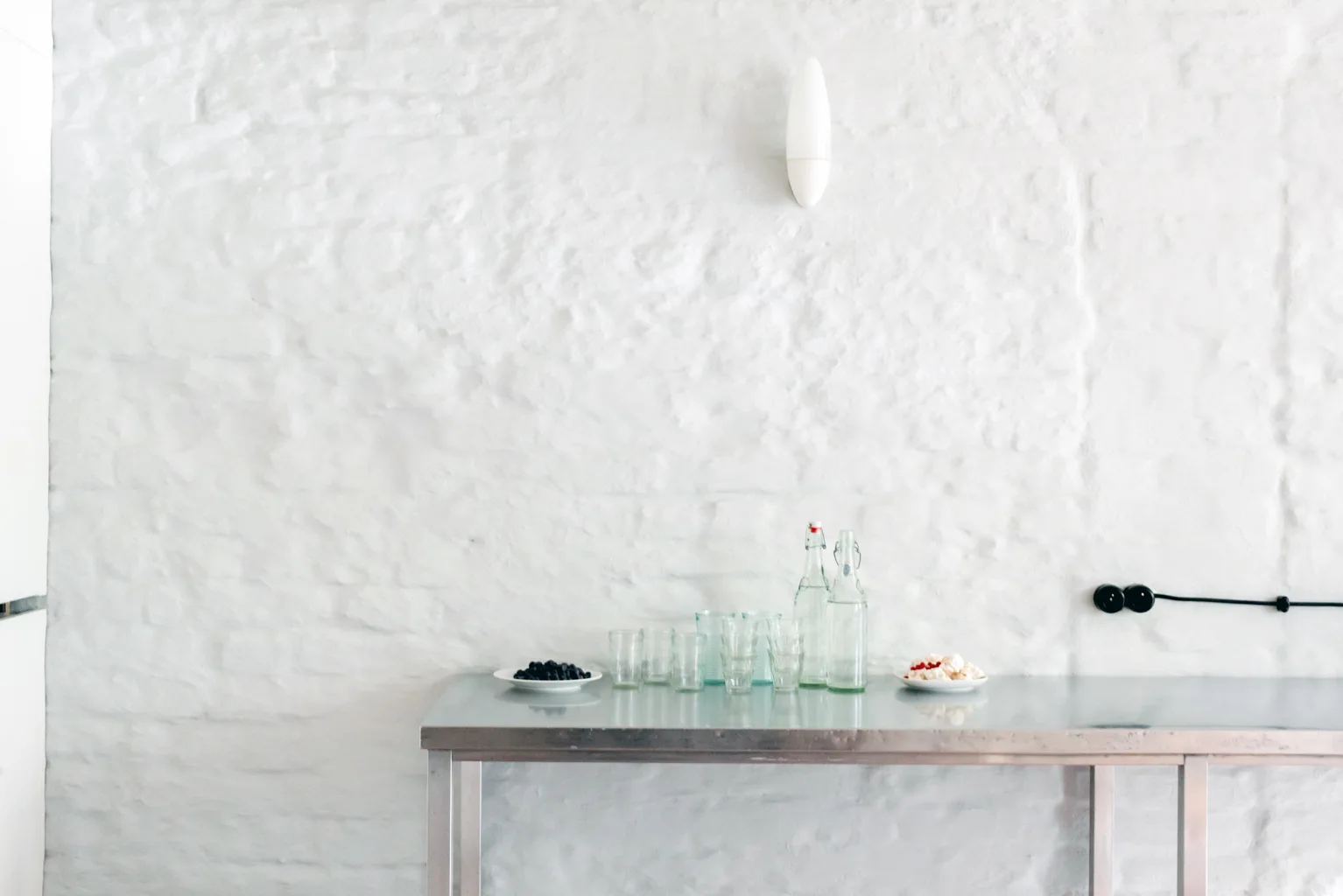 A stainless steel kitchen table stands against a textured white brick wall with a minimalist white wall sconce above it.