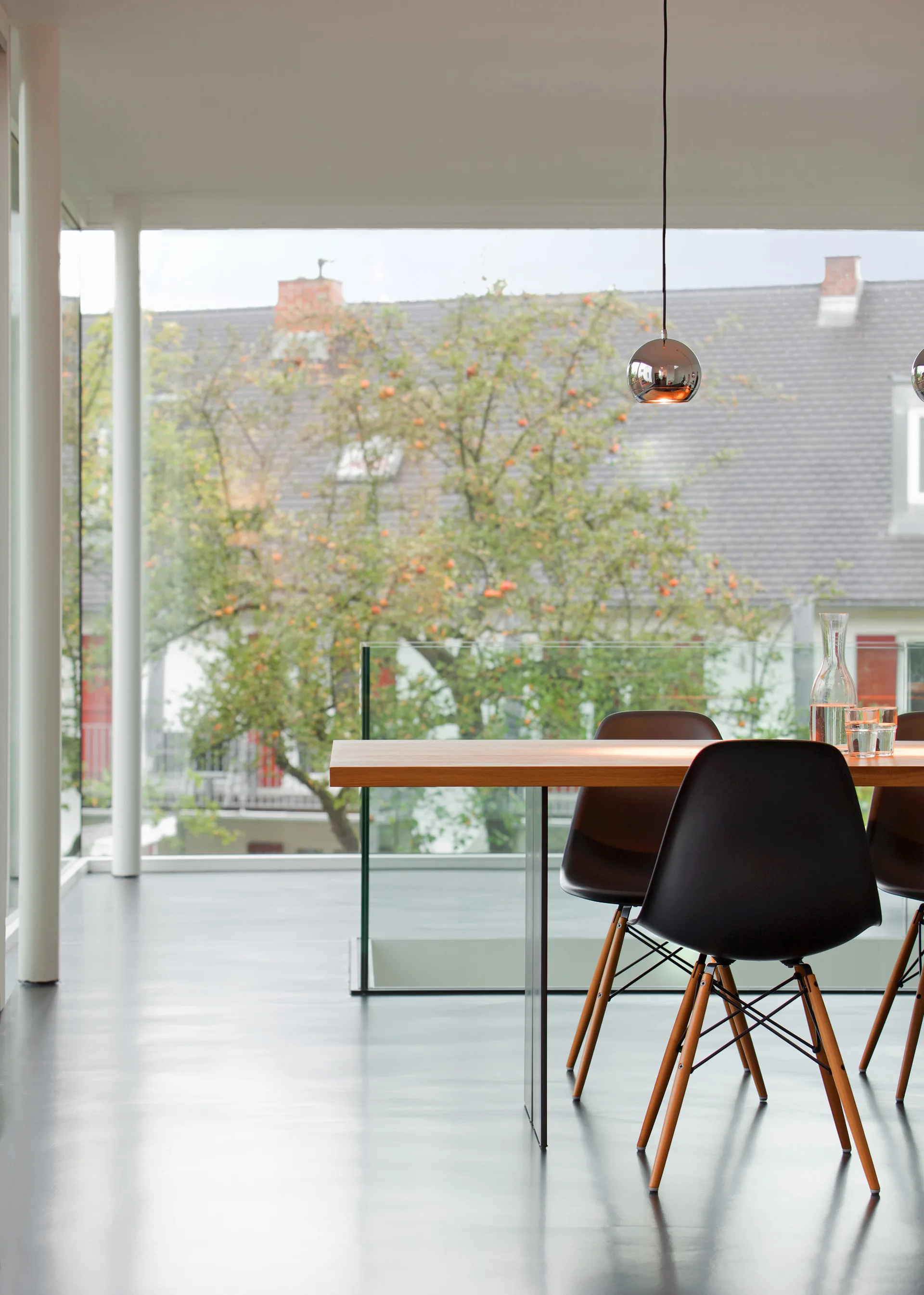 A modern dining room features a wood-topped table and black molded chairs next to a large floor-to-ceiling glass wall.