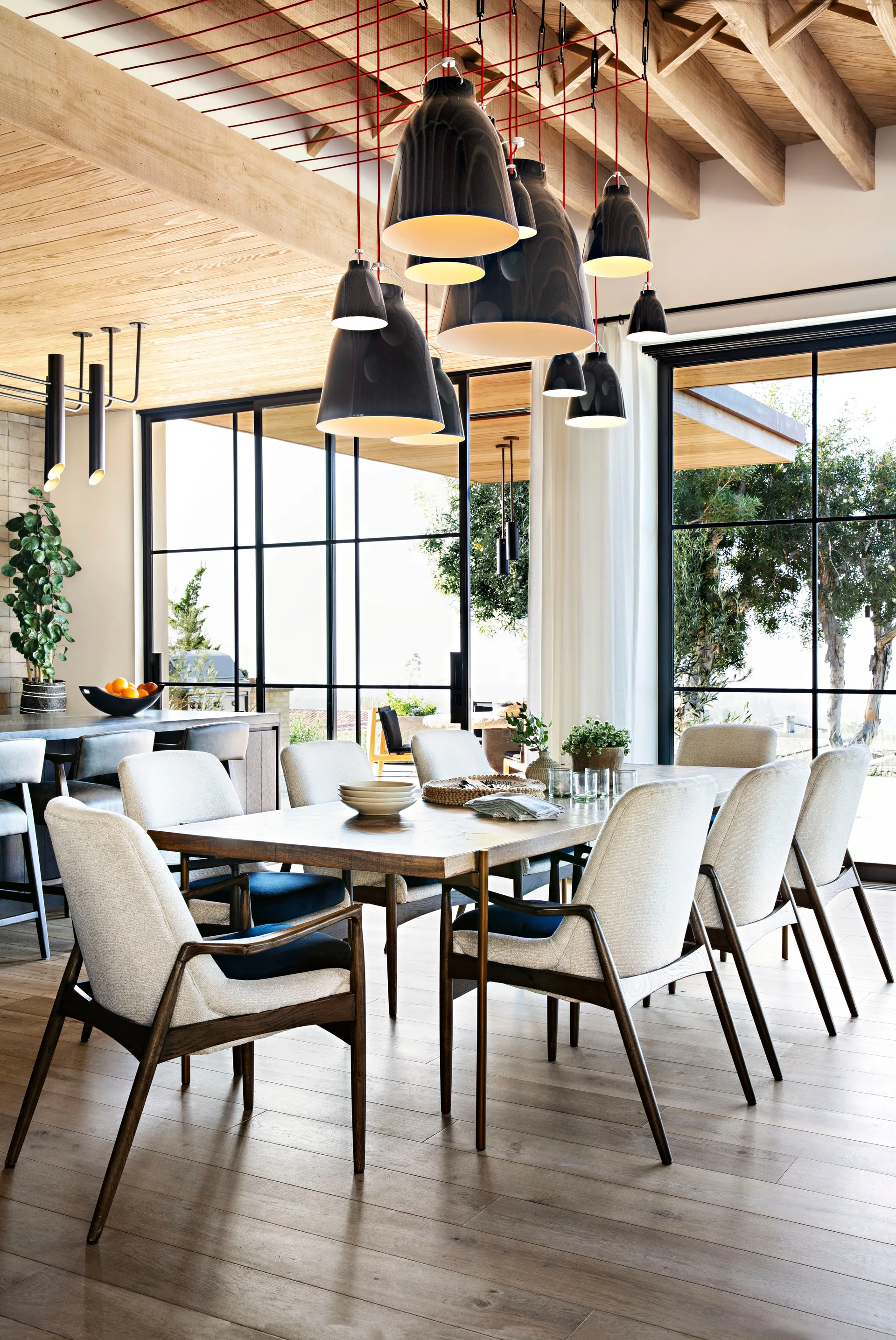 A dining room features a wooden table and upholstered chairs underneath a cluster of black pendant lights hanging from an exposed beam ceiling.