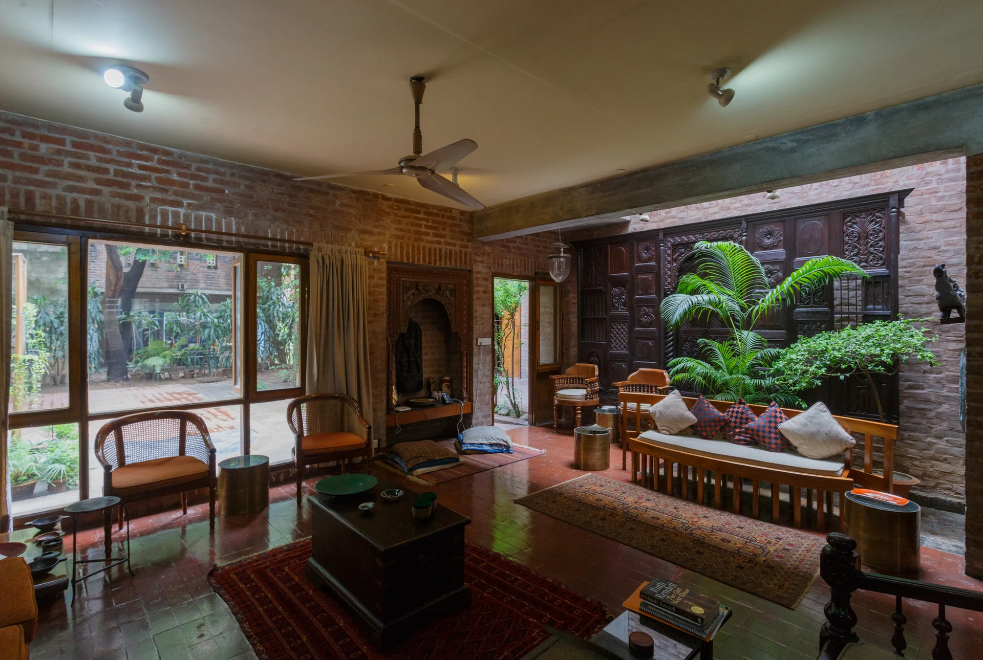 A rustic living room featuring exposed red brick walls, glossy terracotta floor tiles, a carved dark wood wall panel, and traditional wooden seating.