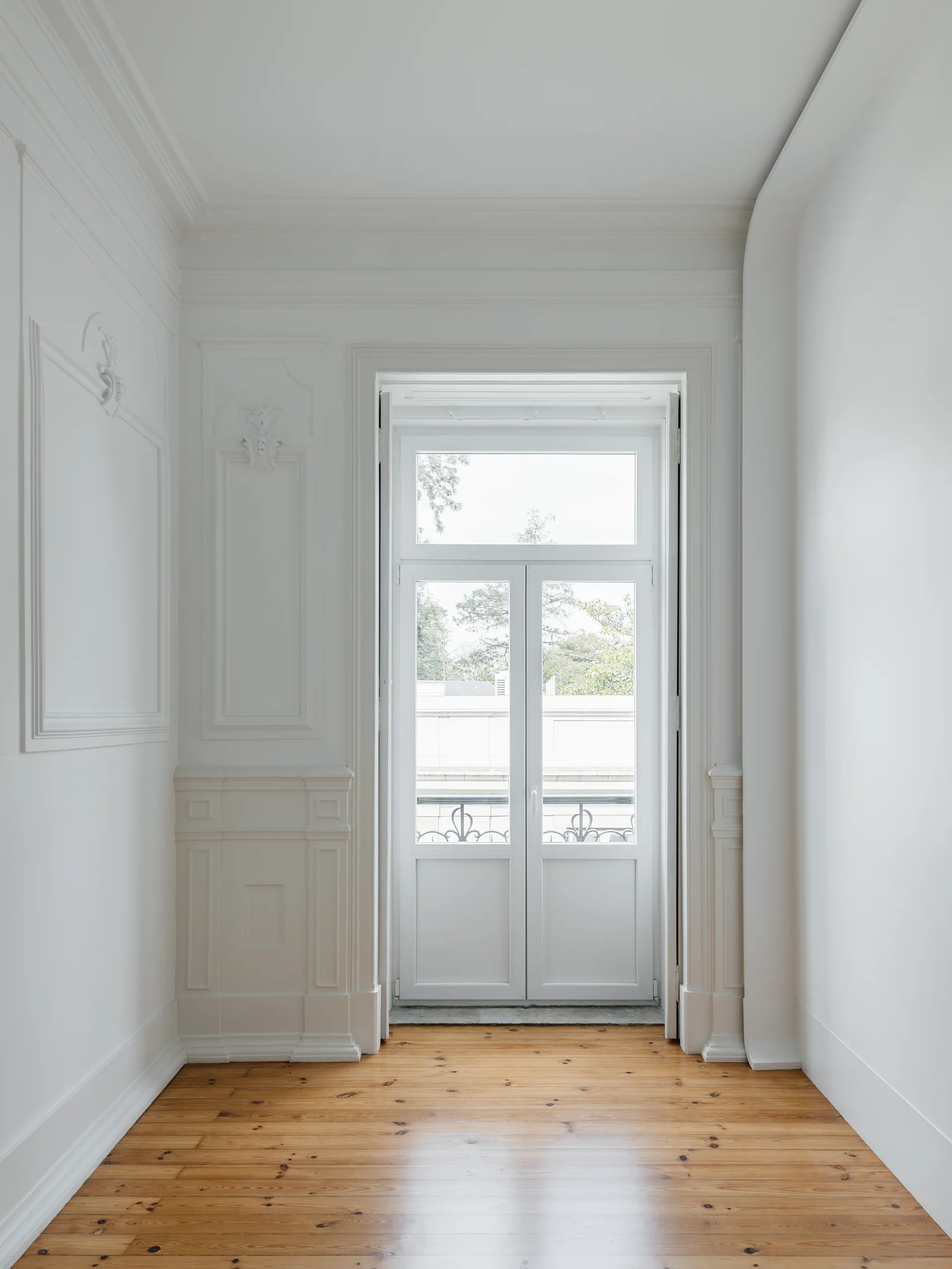 An empty white room featuring traditional wall molding and wainscoting, polished hardwood flooring, and a glass-paned French door with a transom.