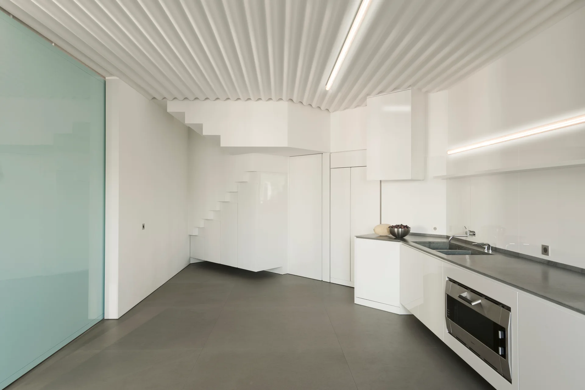 A minimalist white kitchen features integrated cabinets, a grey countertop, a white corrugated ceiling, and a pale blue sliding door panel.