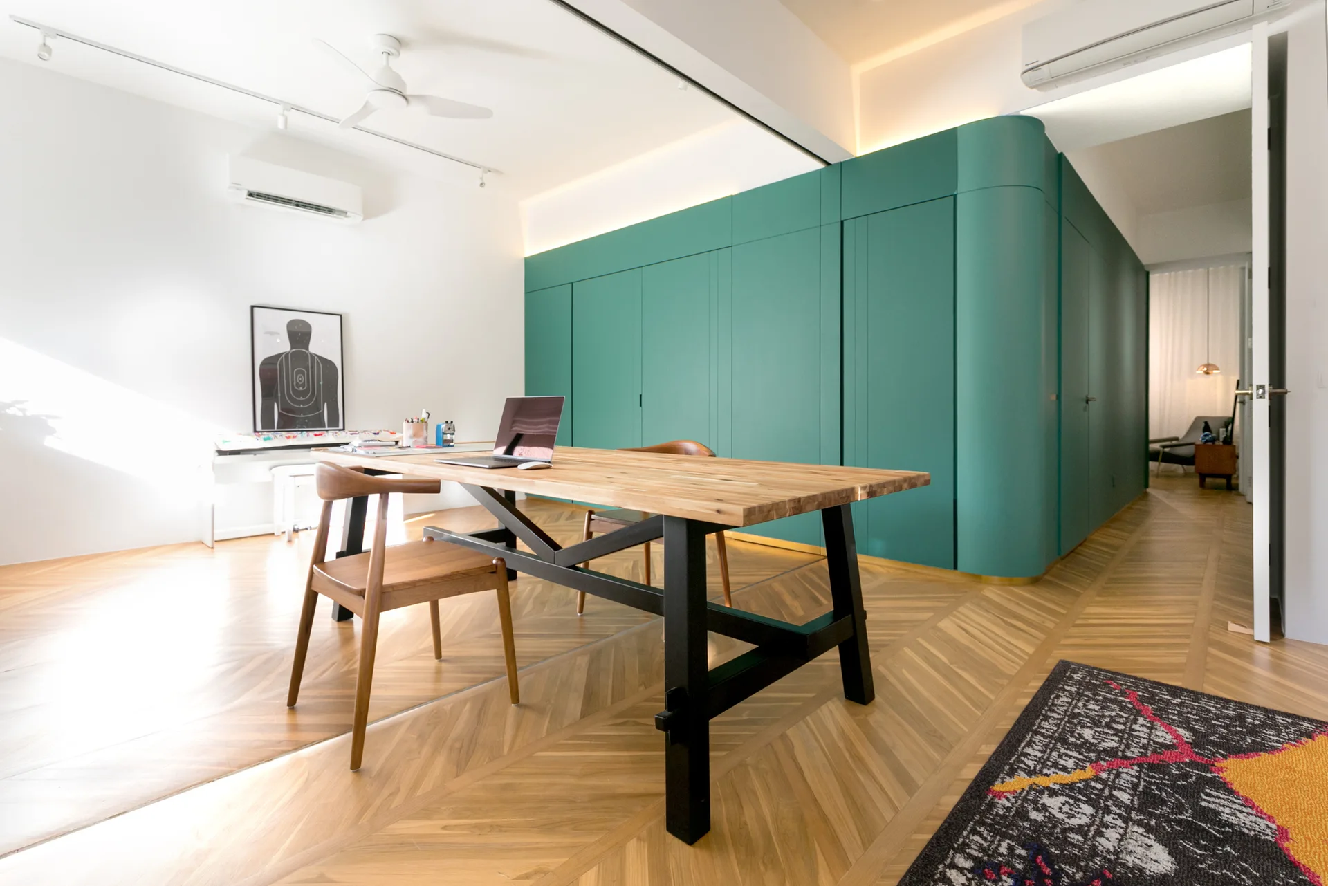 A modern workspace features a wooden desk, wood chairs, a large curved teal storage wall, and herringbone parquet flooring.