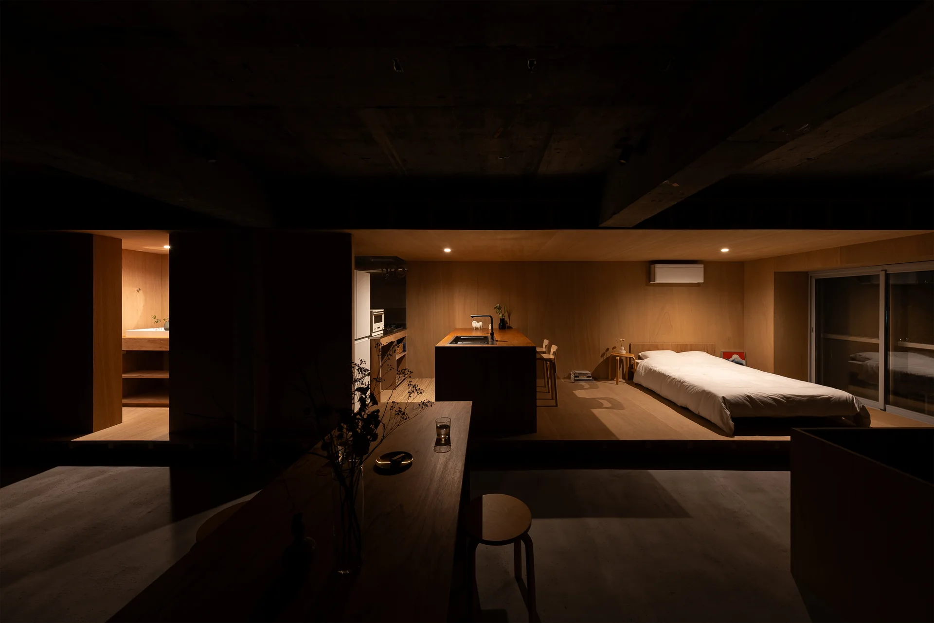 A minimalist studio apartment features a raised wooden platform for a bed next to a kitchen island under an exposed concrete ceiling with warm lighting.
