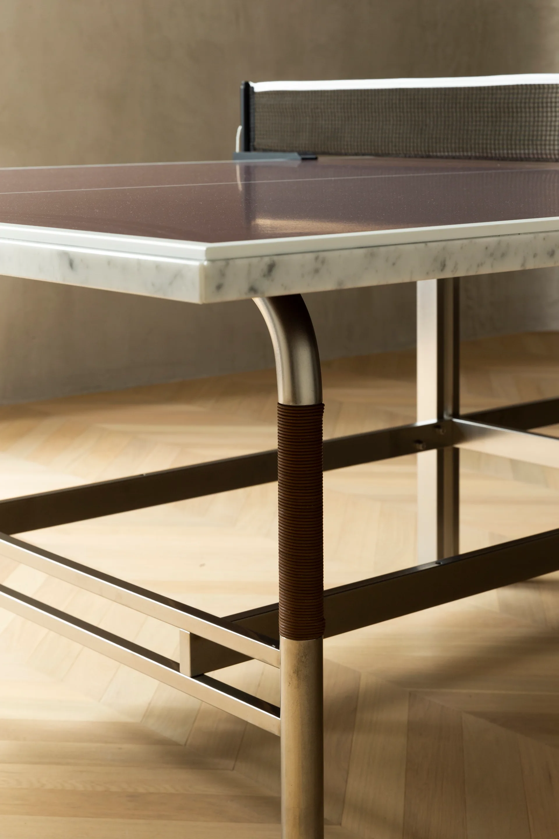 A modern ping pong table features a brown glossy top, a white marble edge, and brushed steel legs wrapped with dark brown leather on a herringbone wood floor.