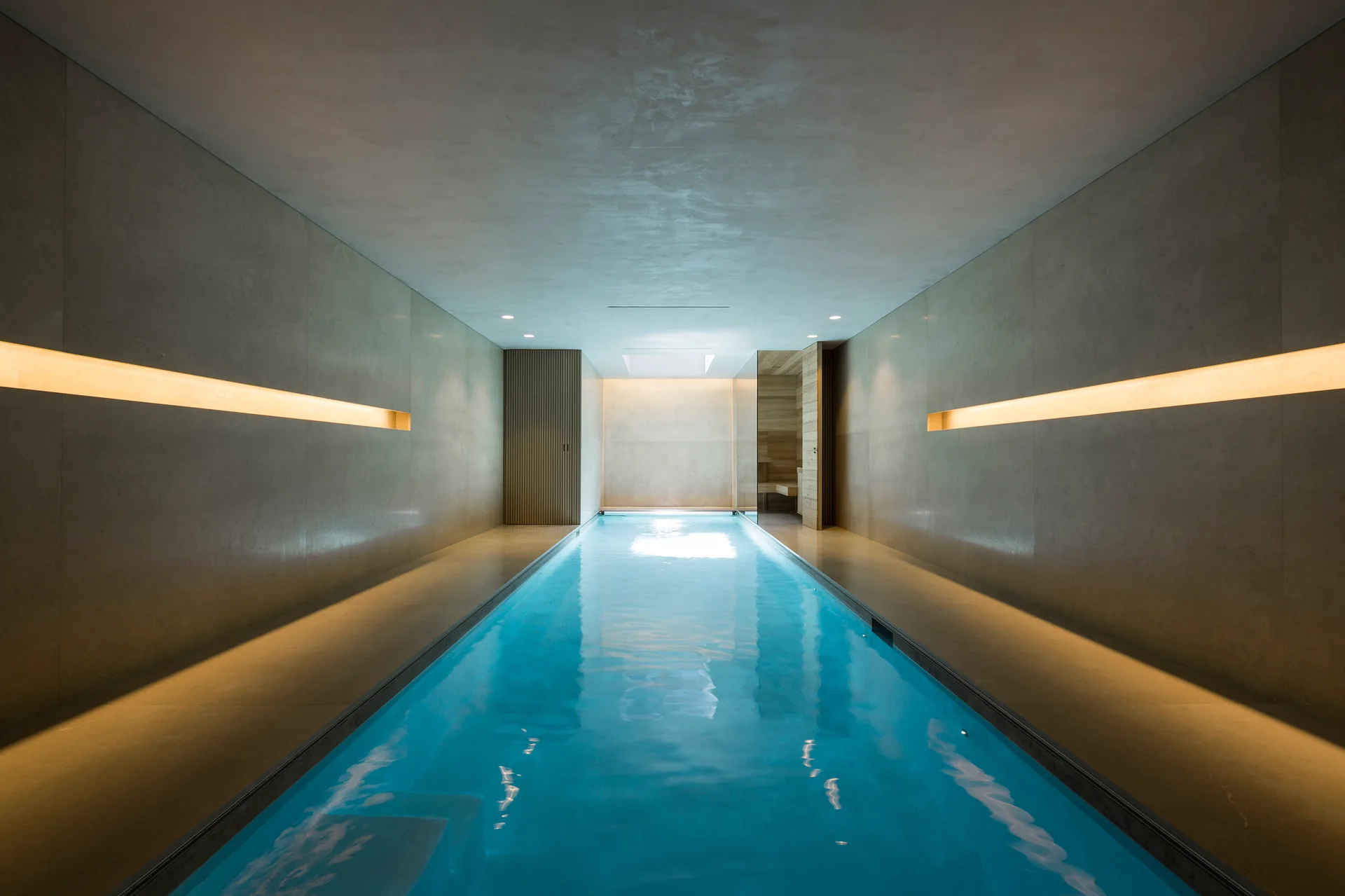 A minimalist indoor lap pool with glowing blue water is bordered by smooth gray stone walls featuring recessed horizontal light strips.