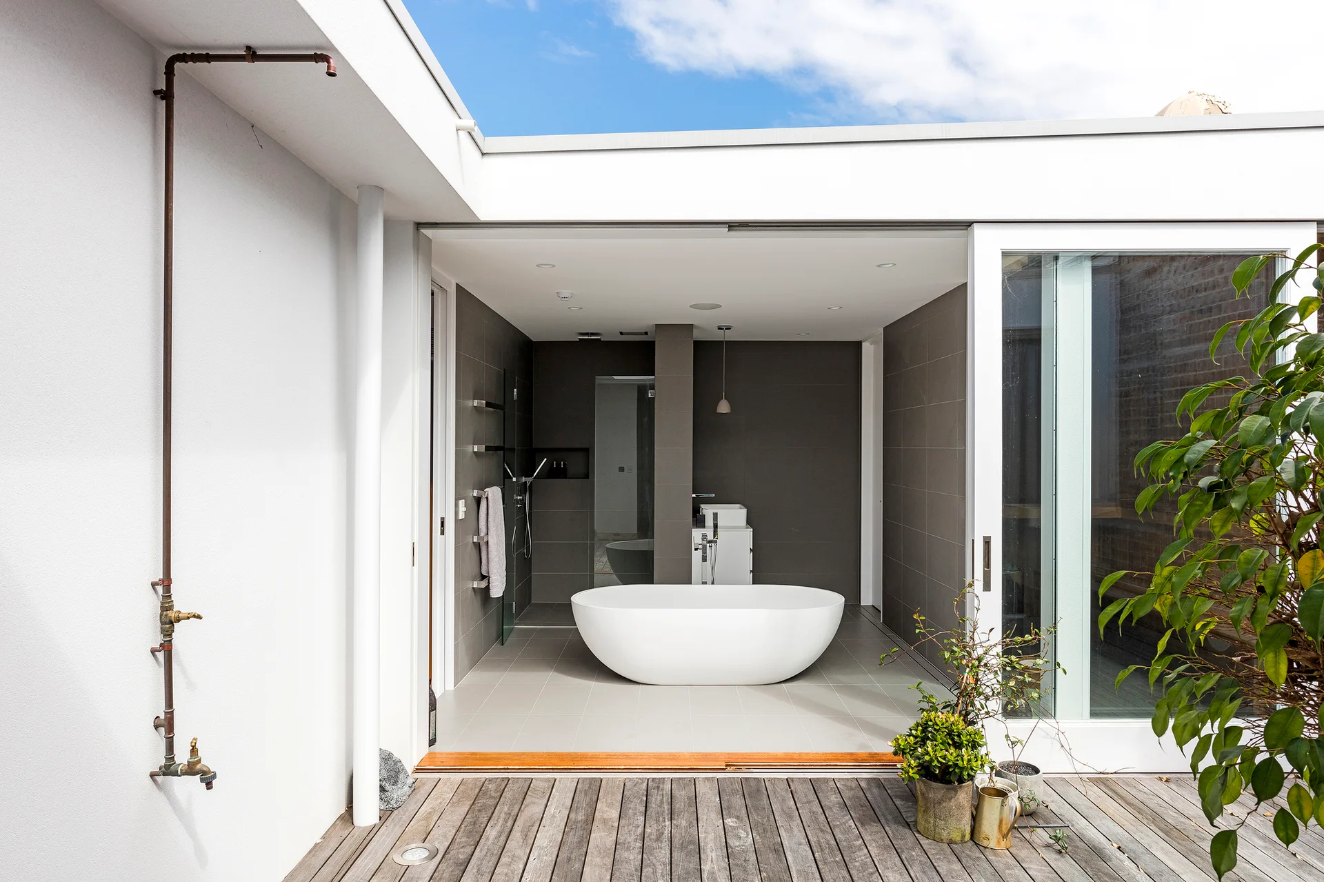 A modern bathroom with a white freestanding bathtub and gray tiled walls opens onto a weathered wooden deck with copper pipes.
