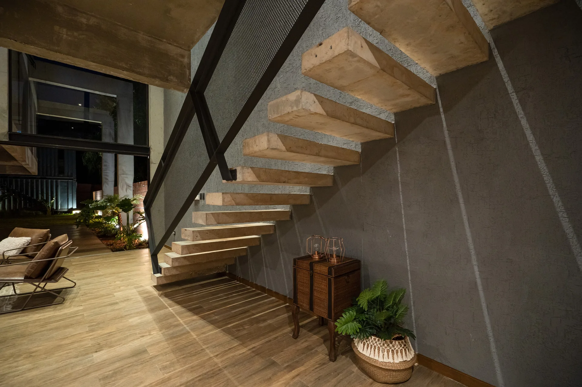 A floating concrete staircase with a black mesh railing climbs alongside a textured grey wall in a modern room with wood floors.