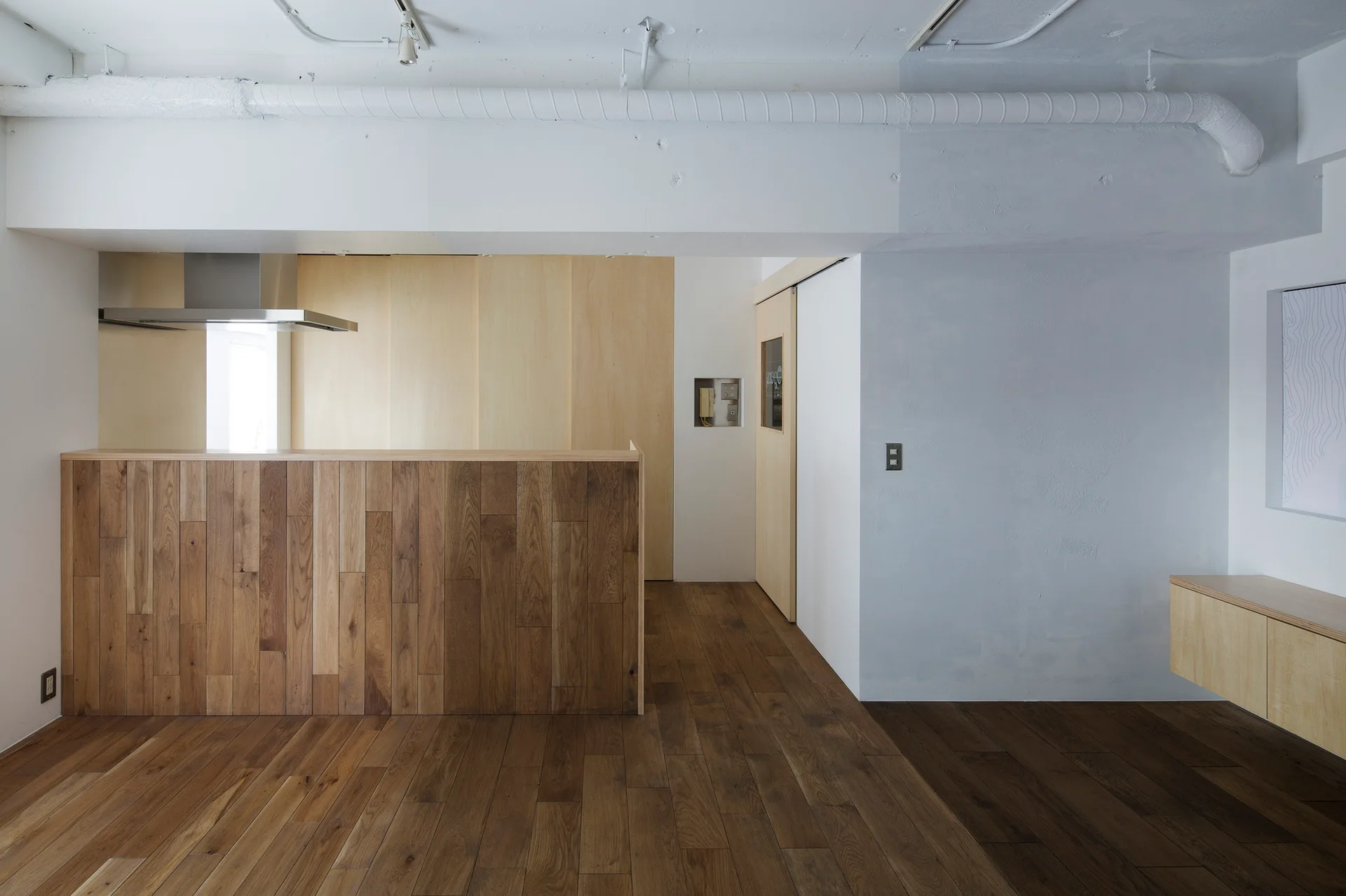 A minimalist kitchen features a kitchen island clad in vertical wood planks, matching dark wood floors, white walls, and an exposed white ceiling duct.