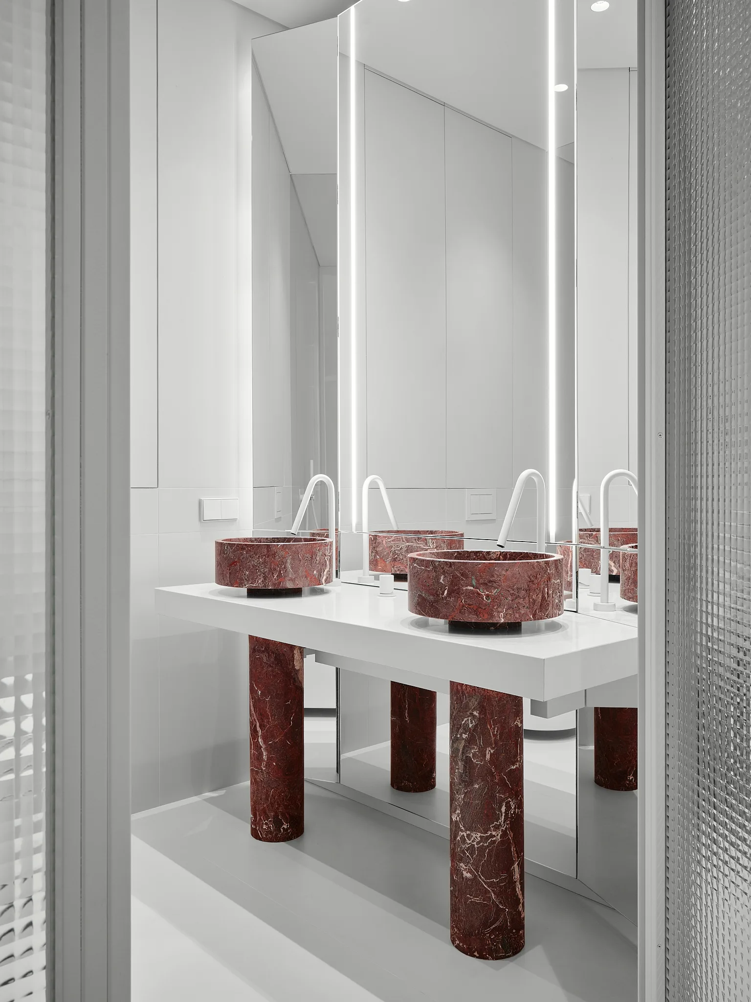 A white bathroom vanity supported by red marble cylindrical legs features matching red marble vessel sinks and white arched faucets.
