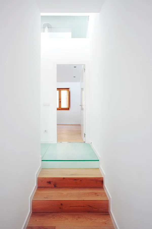 A minimalist staircase features natural wood steps leading up to a translucent glass landing in a bright corridor with white walls.